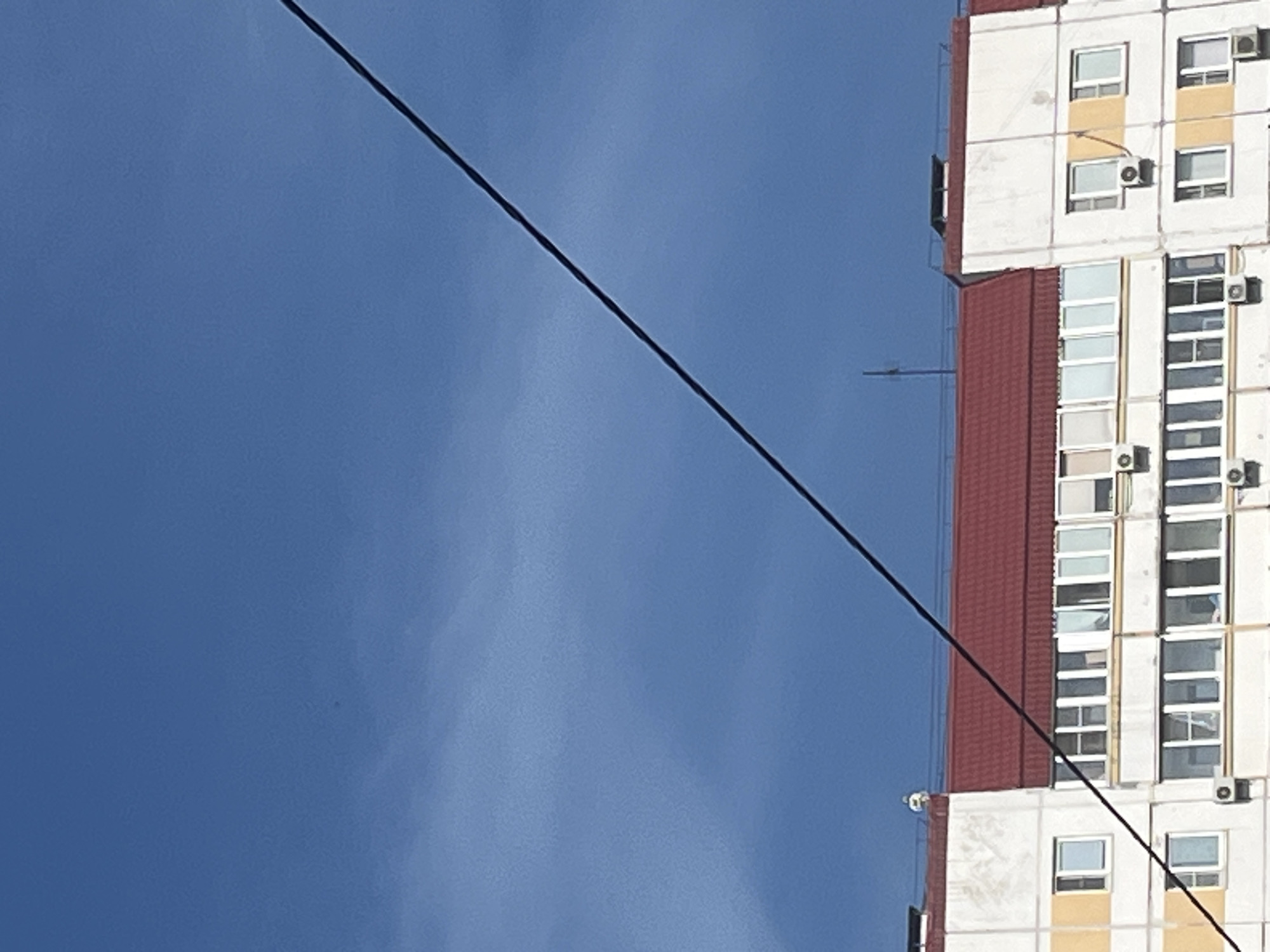 The image displays an outdoor daytime scene dominated by a clear blue sky, a diagonal utility cable, and the upper section of a multi-story building, located in the city of Unknown, Unknown. The sky occupies approximately two-thirds of the upper frame, exhibiting a consistent blue hue with subtle, wispy cloud formations visible, particularly in the lower sky segment. A thick, dark, linear cable traverses the image diagonally from the upper right to the lower left, obscuring parts of the sky and the building.
The visible portion of the building features a prominent red-tiled roof. On the left section of the roof, a small, white, spherical object or structure is present. Towards the center-right of the roof, a thin, black, vertical antenna-like structure protrudes. Further to the right, another rectangular vent structure is visible on the roofline. The building's facade is primarily white with horizontal yellow bands on some levels. Numerous rectangular windows are uniformly distributed across the visible floors. Multiple outdoor air conditioner compressor units are mounted below various windows on the building's exterior walls.