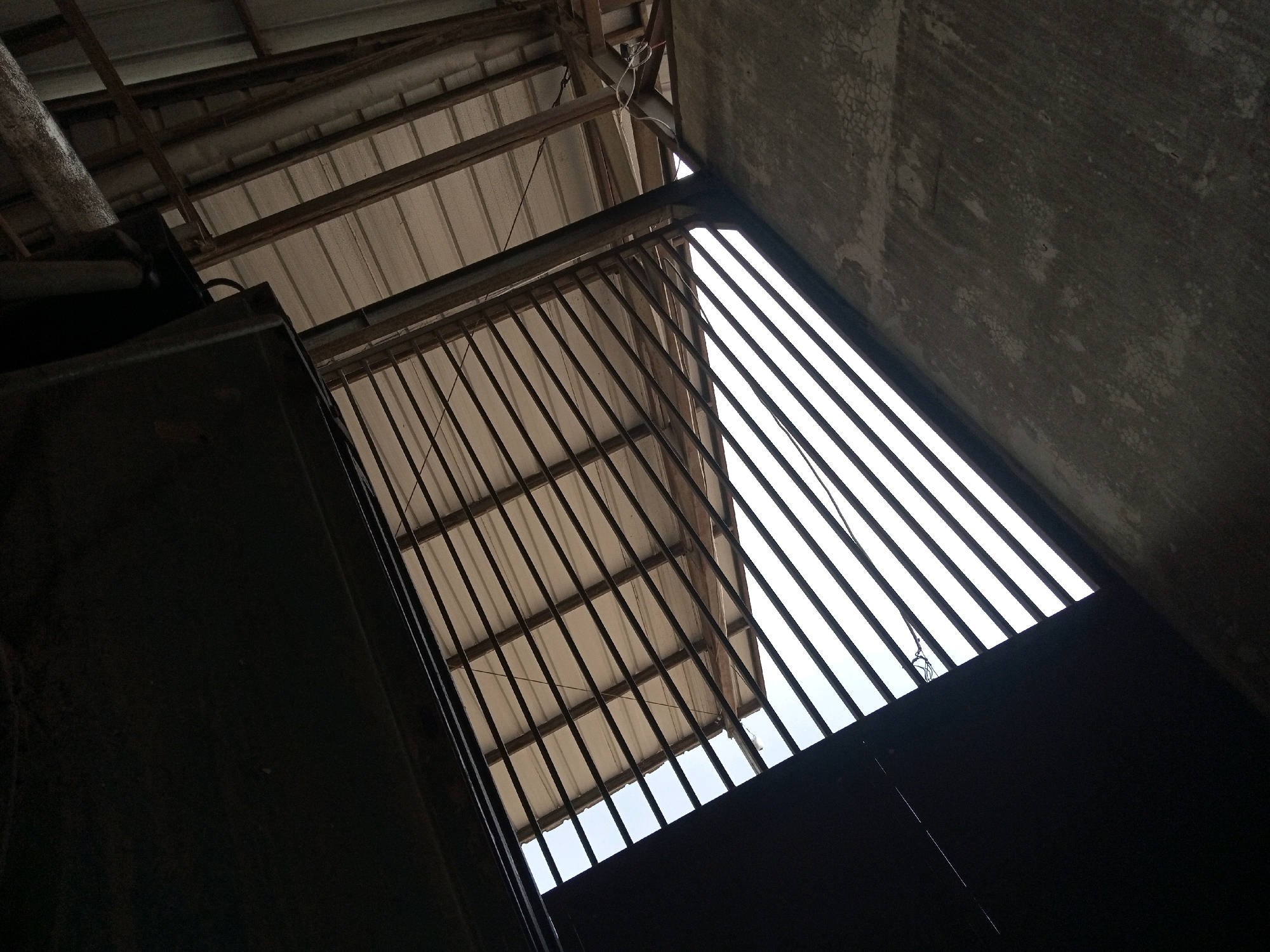 The image depicts a low-angle view looking upwards towards a partially constructed or open-roofed structure. The composition is dominated by geometric lines and shapes formed by roofing materials, metal bars, and concrete walls. In the foreground, a dark, solid object, possibly a wall or a large piece of equipment, occupies the left side of the frame. Extending from this to the right is a dark, slatted structure, resembling a gate or a large window grill.
Beyond the slats, the roof structure is visible, composed of corrugated metal panels supported by wooden beams and possibly some metal framing. The sky or a brightly lit area is visible through the gaps in the slats and under the roofing. The upper right portion of the image features a rough, textured concrete surface, likely a wall or the underside of a concrete structure, showing signs of wear and possibly cracking. The overall impression is of an industrial or utilitarian space, possibly under construction or in a state of disrepair. The lighting suggests it is daytime, with the bright sky providing a strong contrast to the shadowed areas of the structure. The image does not contain any visible people or specific identifiable landmarks that would directly indicate the location of Teluk Dalem Ilir, Indonesia.