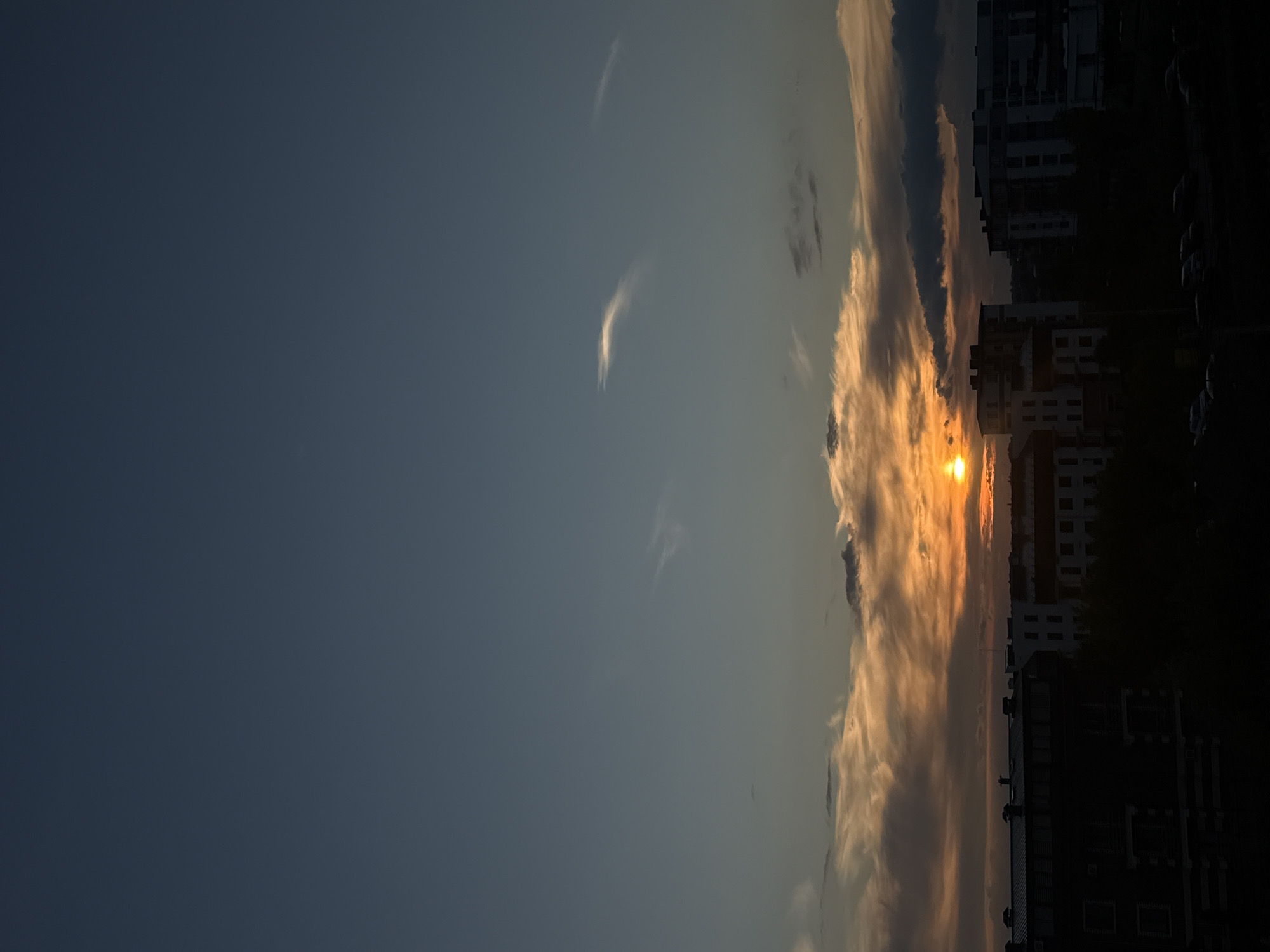 A vertical, eye-level shot captures a city skyline against a dramatic sunset. The sky dominates the upper two-thirds of the frame, transitioning from a deep, dusky blue at the top to warmer, orange and yellow hues near the horizon. Wispy clouds, tinged with the colors of the setting sun, are scattered across the mid-sky.
The lower third of the image is filled with the silhouettes of buildings and trees. In the foreground, a dark building with multiple windows and visible rooftop structures occupies the left side. To its right, several other buildings of varying heights and architectural styles are visible, some appearing to be modern apartment complexes. The setting sun, a bright orange orb, is partially obscured by clouds and appears just above the buildings, casting a warm glow. Dark trees are interspersed between the buildings, further adding to the shadowed silhouette effect. The very bottom of the image shows a dark area that may be a street or parking lot with the faint outlines of several parked cars. The overall scene depicts a twilight urban landscape bathed in the warm light of a setting sun.