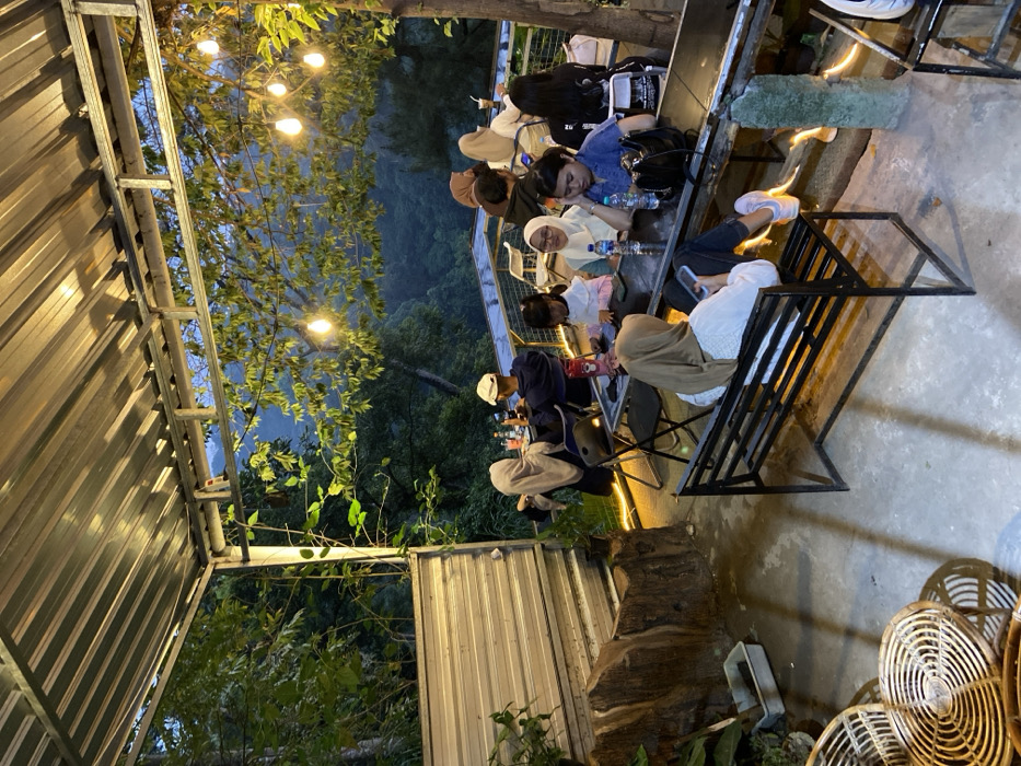The image depicts an outdoor dining or cafe area in Cembor, Indonesia, at dusk or evening, illuminated by overhead string lights. The establishment features a corrugated metal roof supported by a metal frame, overlooking a deep, forested valley with distant mountains under a twilight sky. A metal railing with transparent panels separates the seating area from the natural drop-off.

Approximately twelve individuals are present, consisting of adults and several children, primarily seated at dark rectangular tables with black folding chairs. In the immediate foreground, a person wearing a light brown hijab and white long-sleeved top is seated on a black metal bench, facing away from the camera, engaged with a smartphone.

At a central table, five individuals are gathered. This group includes a young girl in a white long-sleeved top looking at a phone on the table, an adult woman in a light-colored hijab and glasses looking towards the left, and another adult woman in a blue top looking downwards, with a black bag on the table. Water bottles and other beverages are present on this table. Two more individuals are partially visible at this table.

Further into the scene, a smaller table to the left is occupied by two individuals: one in a light-colored hijab and another wearing a white cap and a dark blue long-sleeved top. To the right, a third table is occupied by three individuals, two of whom are wearing hijabs, with one facing away from the camera.

The floor is a light-colored paved surface. In the lower-left, two woven rattan chairs are visible. A large, gnarled, dark brown structure, possibly a decorative tree trunk or sculpted element, stands beside a corrugated metal wall on the far left. Lush green foliage surrounds the dining area, with trees growing within and around the structure.