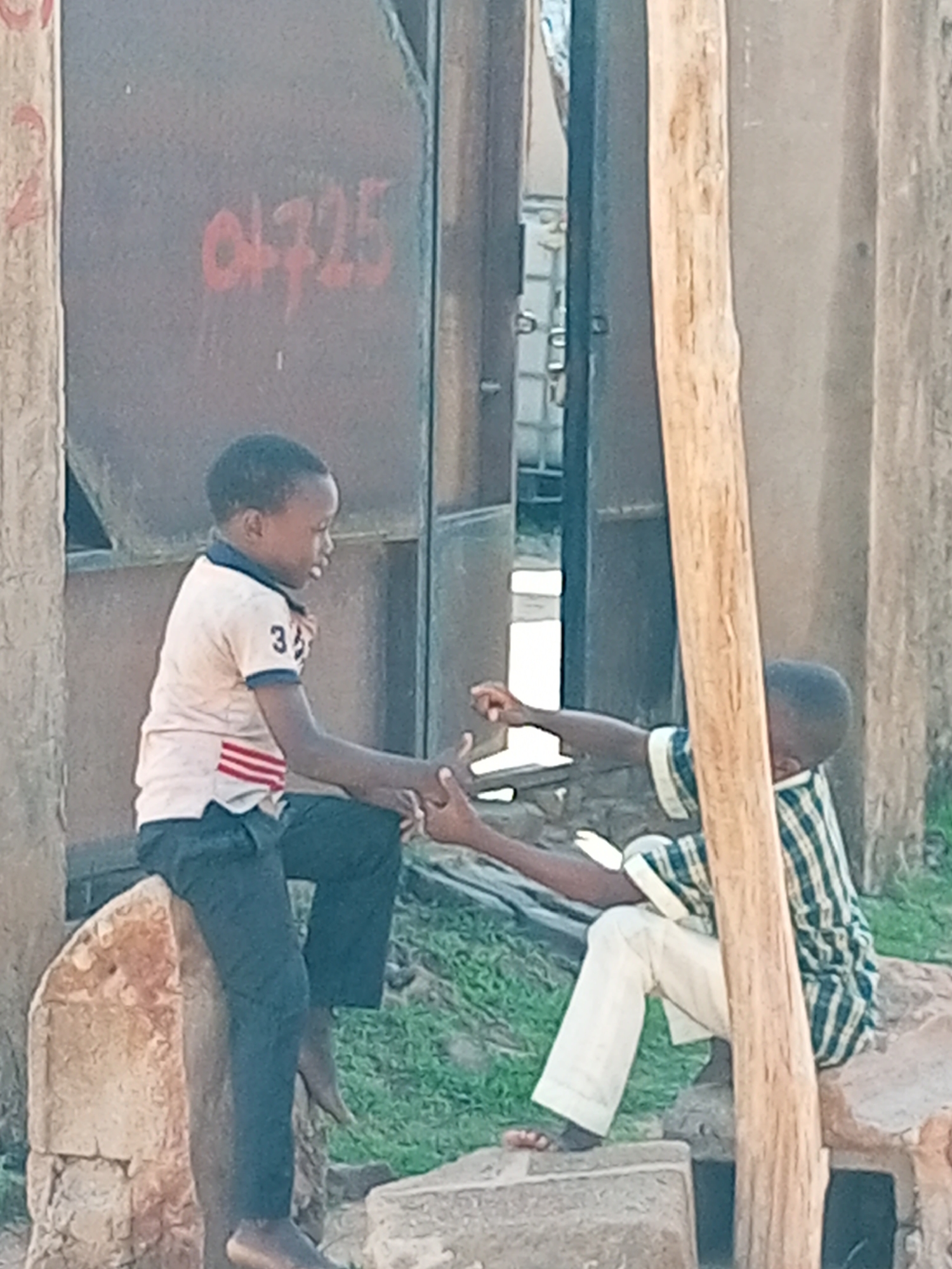 Two young boys are sitting on rocks outdoors. The boy on the left, wearing a light-colored t-shirt with red and blue stripes on the sleeves and dark pants, is shaking hands with the boy on the right, who is wearing a green and white striped shirt and light-colored pants. They are in a dusty environment with a metal structure in the background. The metal structure has red markings that appear to be "04725". The setting suggests a possibly informal settlement or a less developed area within the city of Gombe, Nigeria, possibly near a market or construction site, given the presence of building materials and the general appearance of the surroundings. The lighting suggests it is daytime, possibly late morning or afternoon.
