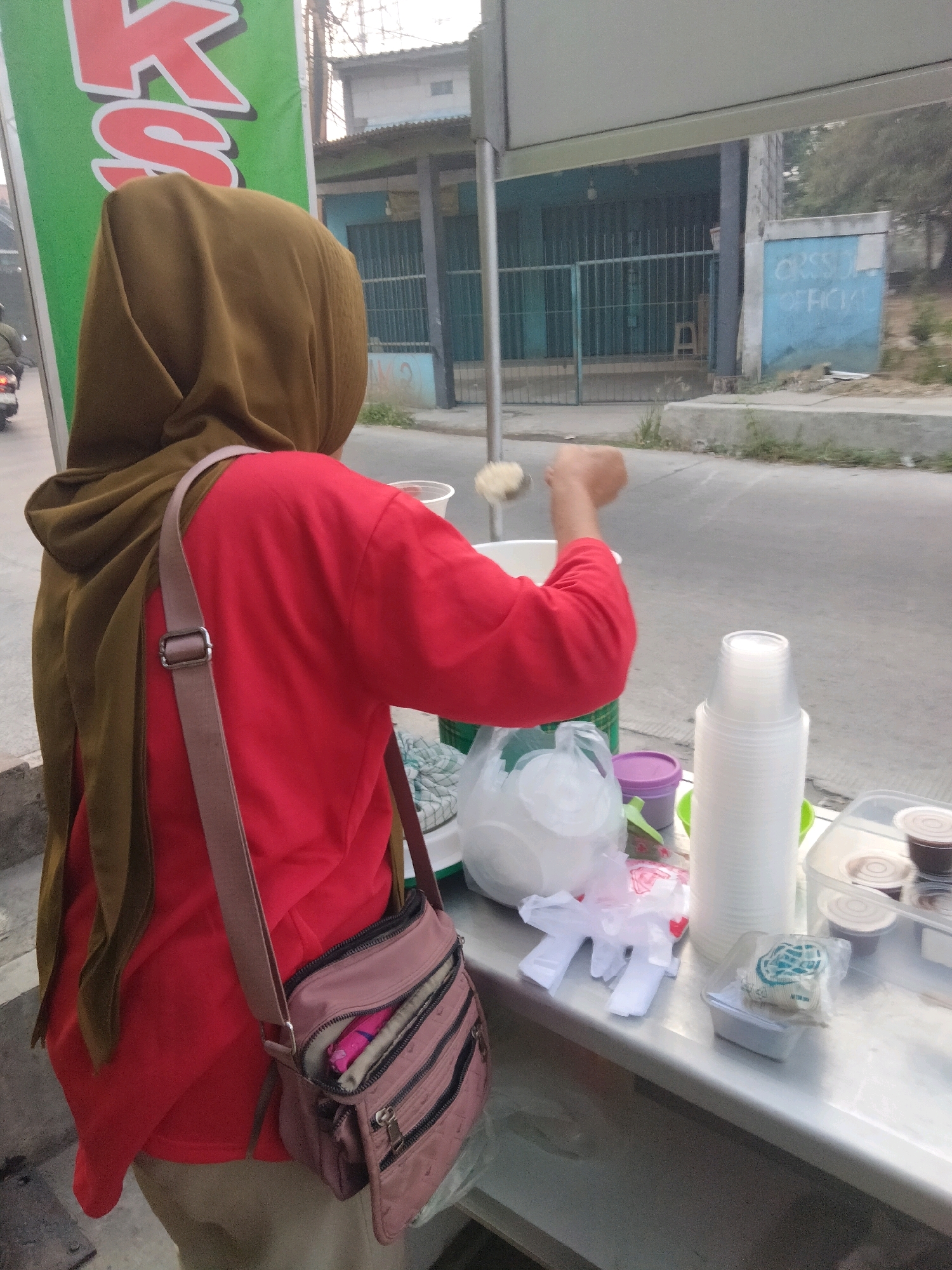 A woman wearing a brown hijab and a red shirt stands behind a food stall on the street in Pisangan Jaya, Indonesia. She is holding a ladle in her right hand, scooping something from a large white container. A pink crossbody bag is slung over her shoulder. On the stall's counter, there are stacks of plastic cups, small containers of sauce or toppings, plastic gloves, and other food service items. In the background, a green banner with red lettering is visible. Beyond the stall, a road and buildings with barred windows can be seen.