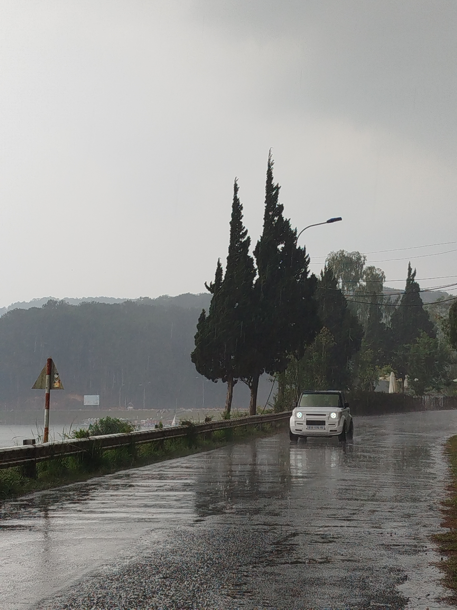 A white SUV with the license plate "61K-176.15" drives on a wet road in Da Lat, Vietnam. Rain is falling, obscuring the view of a lake and forested hills behind the vehicle. A guardrail lines the left side of the road, with sparse vegetation growing alongside it. To the right of the vehicle, there are trees and a lamppost. A road sign is visible on the left side of the road.