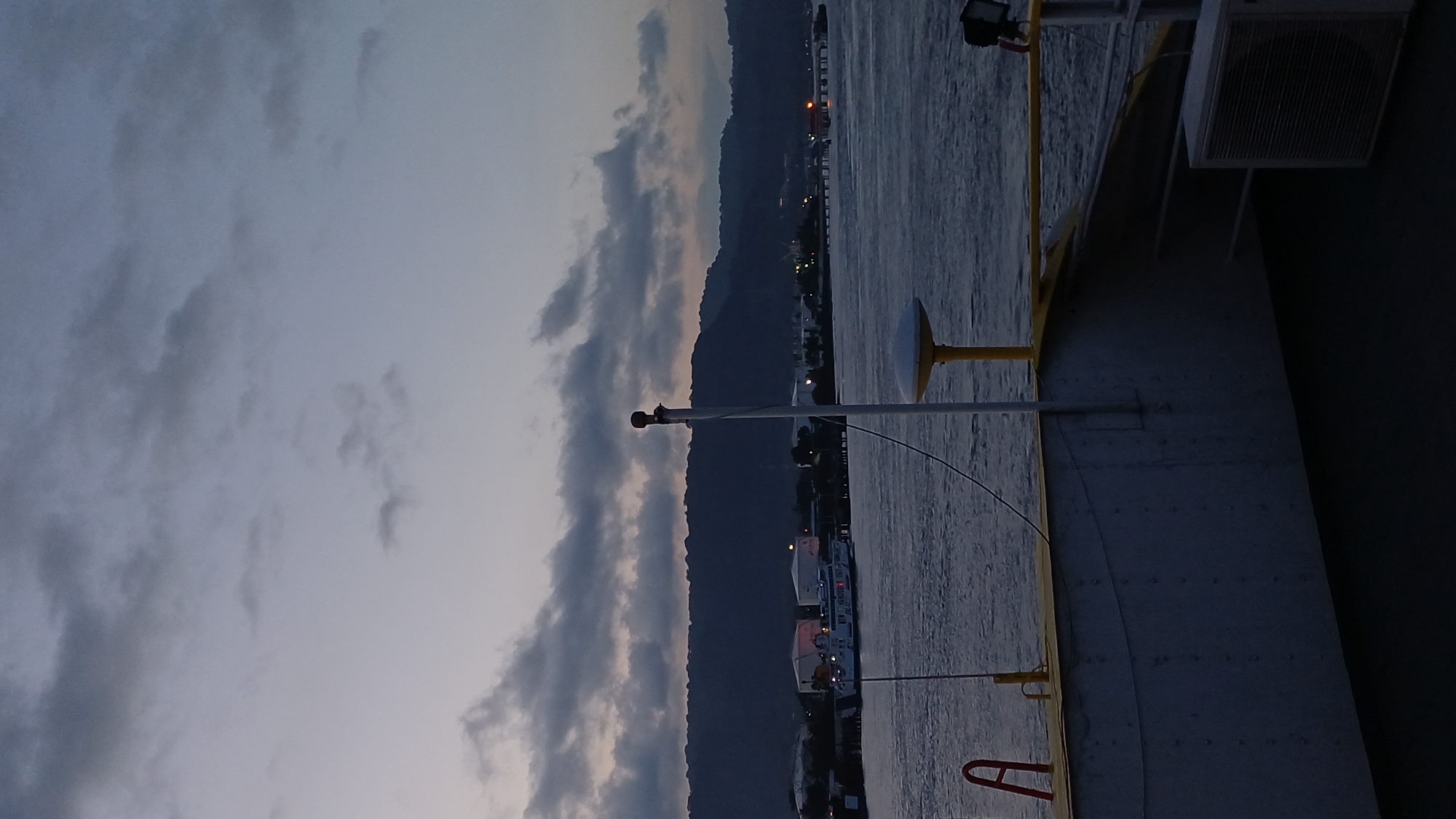 The image depicts a view from a ship towards a city at dusk. In the background, a mountain range stretches along the horizon, its silhouette contrasting with the partially cloudy sky. The city's lights are visible along the coastline, suggesting a settled area. The foreground is dominated by parts of the ship itself, including a yellow post, what appears to be an antenna or signal device, and an air conditioning unit, all of which are connected to the ship's superstructure. The sea is calm, reflecting the dim light of the sky.