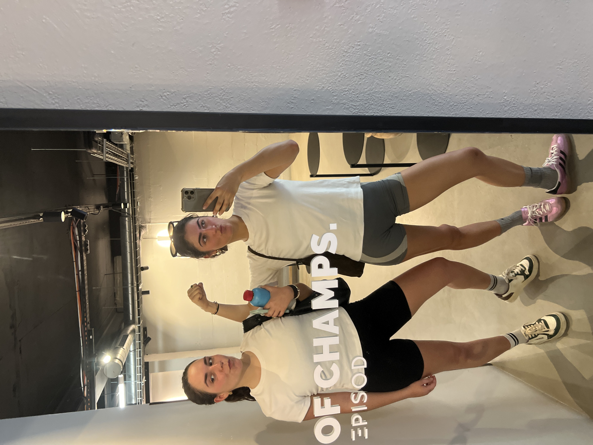 Two young women pose for a mirrored selfie. One woman is holding a phone, and the other is holding a blue water bottle. Both are wearing white t-shirts and athletic shorts. The woman holding the water bottle is wearing black shorts and white tennis shoes. The other woman is wearing gray shorts and pink tennis shoes. The location appears to be an interior with industrial ceiling fixtures. There is an "OF CHAMPS. EPISODE" signage is visible in the mirror's reflection. The city is Paris, France.