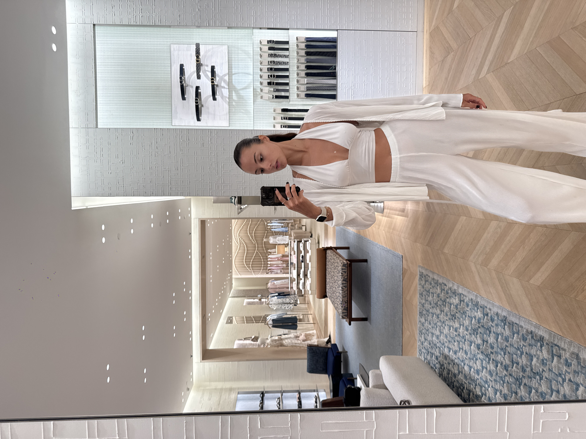 A woman with dark hair stands inside a store, taking a selfie in a mirror. She is wearing a white outfit consisting of a crop top, pants, and a light jacket. The store displays belts and other clothing items in the background. The interior features a patterned wooden floor and a patterned light-colored wall in the foreground. The store appears to be well-lit with white lighting, and there are seating arrangements. The location is Wichit, Thailand.