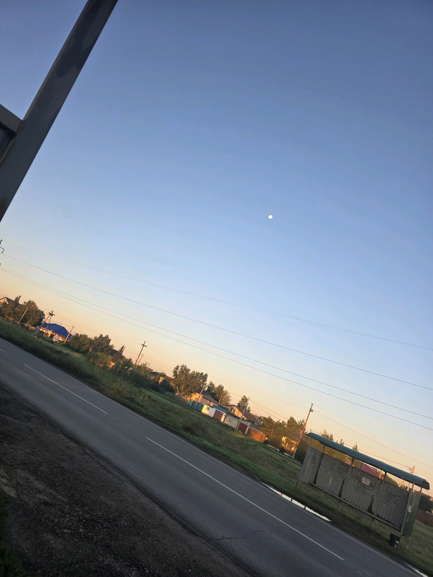 This image depicts a roadside scene at dusk in the vicinity of Славгородское, Russia. The foreground is dominated by a gray asphalt road with white lane markings, angled diagonally across the frame from lower left to upper right. To the left of the road, a dark, textured embankment slopes upward. To the right of the road, a weathered bus stop shelter with a green roof and gray, dilapidated panels is visible. Beyond the road, a grassy verge is dotted with trees and the rooftops of houses, suggesting a rural or suburban setting. The sky is a gradient of pale blue, transitioning to a soft orange hue near the horizon, and a small, pale circle, possibly the moon, is discernible in the upper portion of the sky. Overhead power lines are strung between utility poles, further indicating a developed area. The overall impression is one of a quiet, early evening in a Russian settlement.