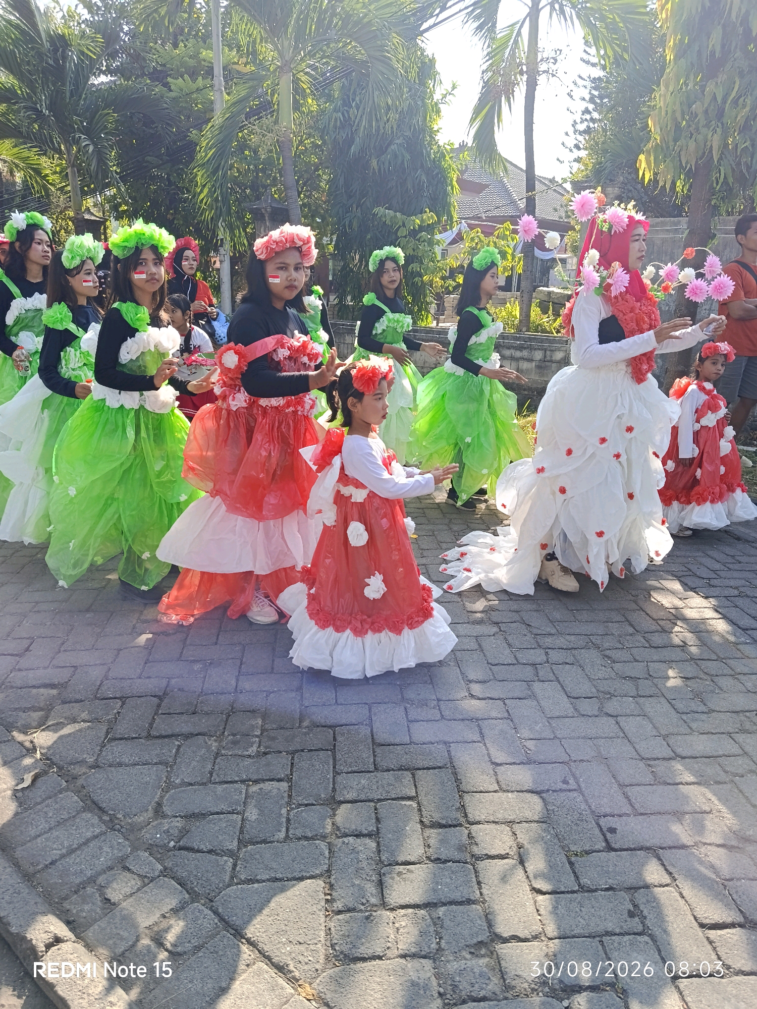 In the city of Keboansikep, Indonesia, a group of young women and a child are participating in a parade. They are dressed in elaborate, colorful costumes that appear to be made of recycled materials, such as plastic bags. The women are wearing flower crowns and are decorated with face paint. The costumes are layered and voluminous, with one woman wearing a white gown adorned with red flower accents and a long train, while others are in vibrant green and red outfits. The child in the foreground is dressed in a red and white costume. They are walking on a paved pathway lined with palm trees and foliage, suggesting an outdoor, tropical setting. The sun is casting shadows on the ground, indicating it is likely daytime.