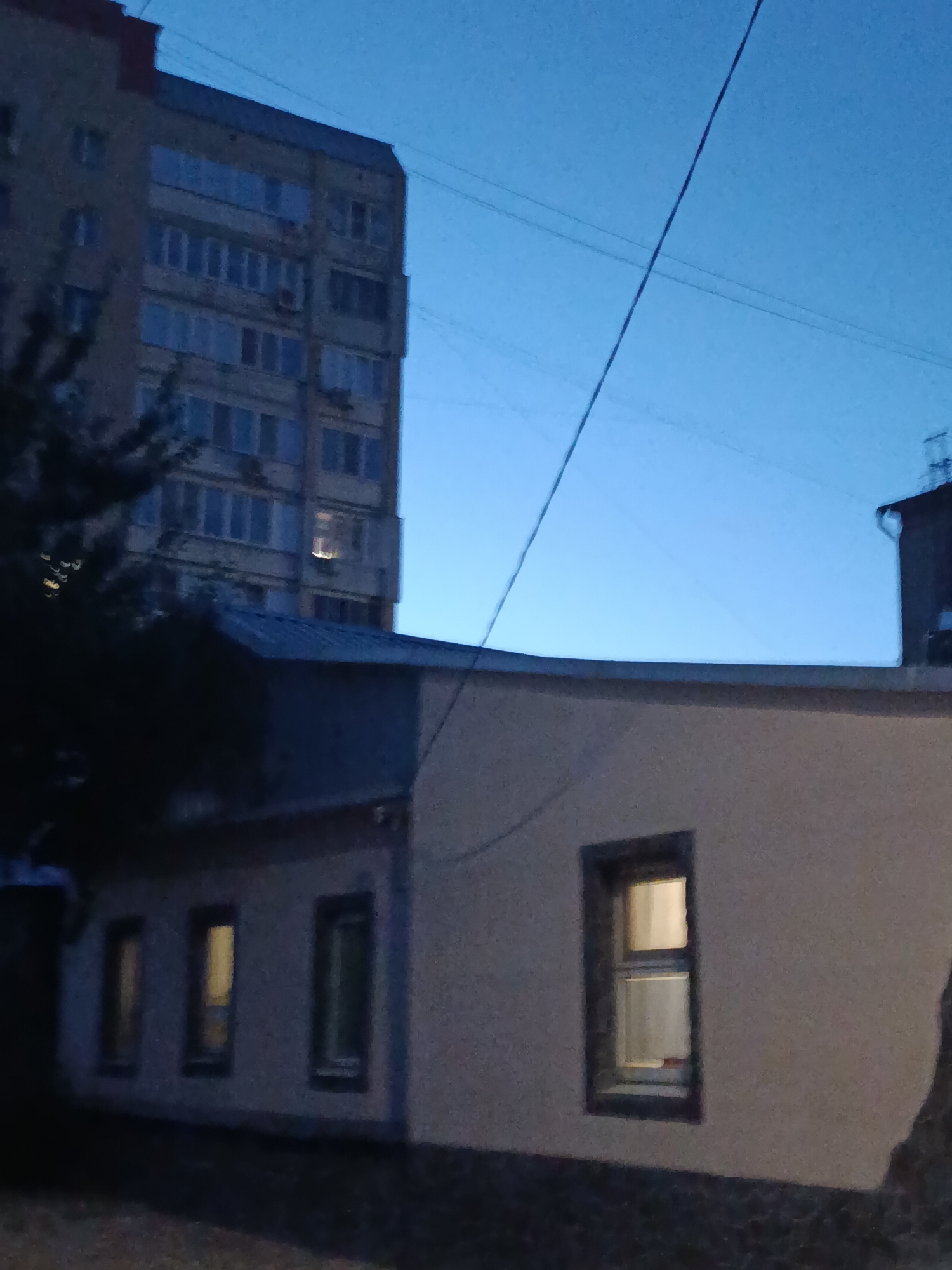 The image depicts an exterior view of buildings at dusk in Saratov, Russia. In the background, a tall, multi-story residential building with numerous windows and balconies is visible against a clear blue sky. In the foreground, a lower, light-colored building with a dark roofline features several windows, some of which are illuminated from within. Power lines crisscross the sky above the structures. The overall scene is dimly lit, suggesting it is either early morning or late evening.