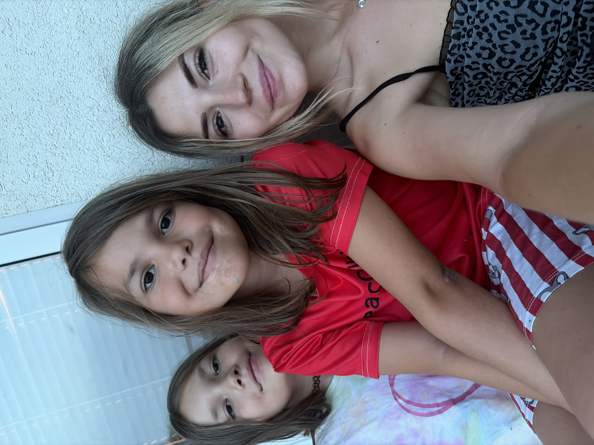 The image shows a close-up, vertical selfie of three individuals, likely two children and one young adult, possibly sisters or friends. They are lying down and looking upwards towards the camera. The lighting suggests it is daytime, possibly outdoors or near a window with natural light.
The person at the top appears to be a young woman with blonde hair, wearing a black spaghetti strap top and a grey and black leopard-print garment. She is smiling.
Below her, a child with brown hair, wearing a red t-shirt with white stitching, is also smiling and looking at the camera. Some light discoloration is visible on her chin and cheek.
At the bottom, another child with brown hair, wearing a tie-dye shirt with pink and white colors, is visible. They have a necklace with a black choker band. This child is also looking at the camera with a slight smile.
The background consists of a textured, light-colored wall on the left and a blurred, reflective surface with horizontal lines on the right, possibly a window or a mirrored surface. The overall mood is casual and friendly. The image appears to be taken in Budva, Montenegro.