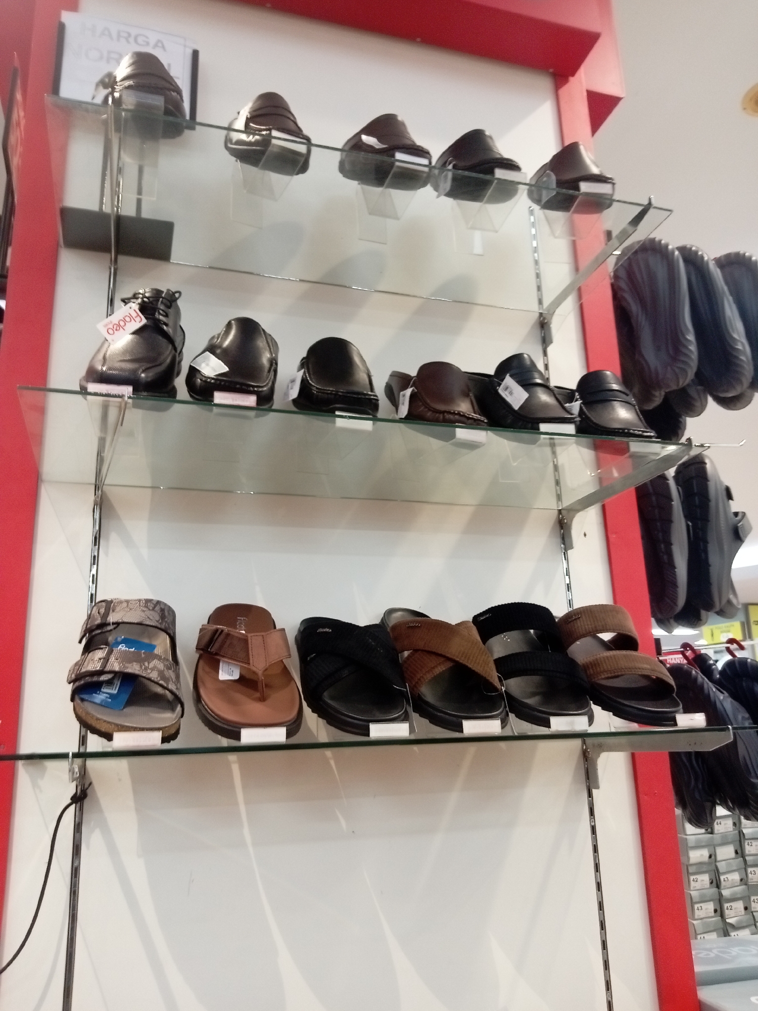 The image displays a retail display of men's footwear on multiple shelves in what appears to be a shoe store. The store is located in Batam, Indonesia, as indicated by the "HARGA NORMAL" sign visible in the top left, which translates to "Normal Price" in Indonesian.
The display features several rows of shoes. The top shelf shows four pairs of dark-colored loafers or dress shoes, each placed on clear acrylic stands. Below this, the second shelf holds six pairs of black leather shoes, also on acrylic stands, with price tags visible on some. The third shelf displays an assortment of sandals and flip-flops. From left to right, there's a pair of patterned sandals with velcro straps, a brown leather flip-flop, a black sandal, a brown cross-strap sandal, and another black sandal with a strap. All these are resting directly on the glass shelf. On the right side of the image, behind the footwear display, several dark-colored shoes are hanging, possibly from a rack. Further to the right, on a lower level, boxes of shoes are stacked, with sizes like "44", "42", and "43" marked on them, suggesting an inventory area.
The environment is brightly lit, typical of a retail setting. The background is mostly white, with red accents from the store's structure. There are no people visible in the frame, making it a static product display. The overall impression is one of an organized retail environment showcasing a variety of men's footwear.