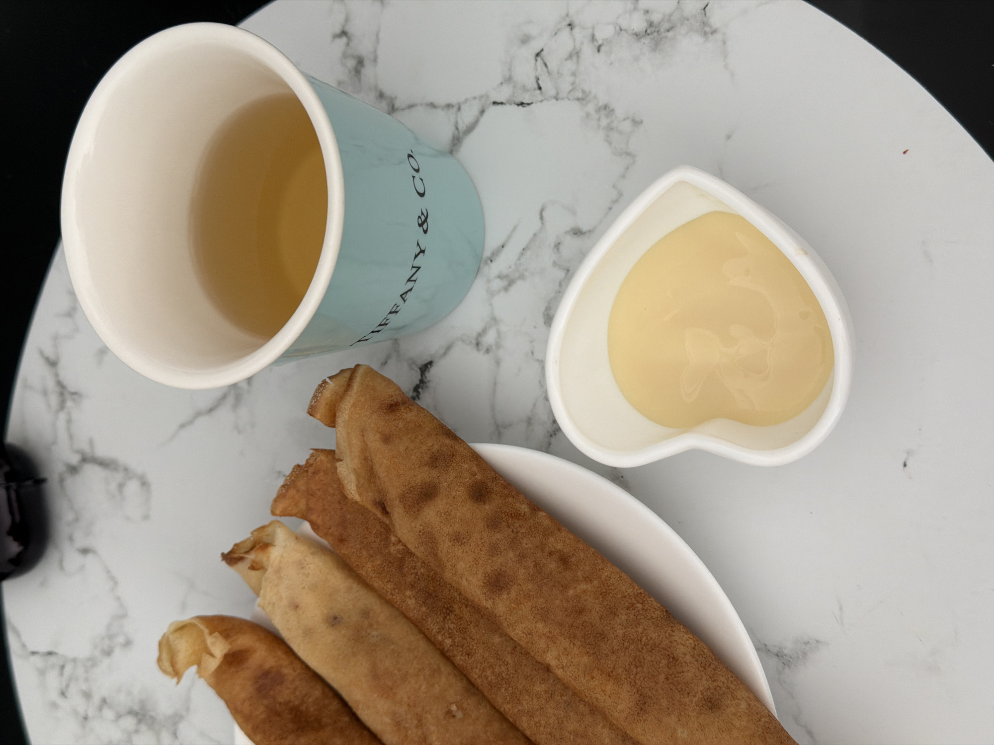 The image shows a close-up overhead view of a breakfast setting on a marble-patterned surface. A light blue Tiffany & Co. mug filled with a golden-colored liquid, likely tea or coffee, is positioned to the right. In front of the mug is a small, white, heart-shaped dish containing a pale yellow, creamy substance, possibly condensed milk or a sweet sauce. To the left, on a white plate, are several rolled crepes or thin pancakes, appearing golden brown and slightly crisp. The overall scene is well-lit, with the focus on the food and drink. There are no people visible in the image, and no specific landmarks or features within Finestrat, Spain, are identifiable from the provided view.