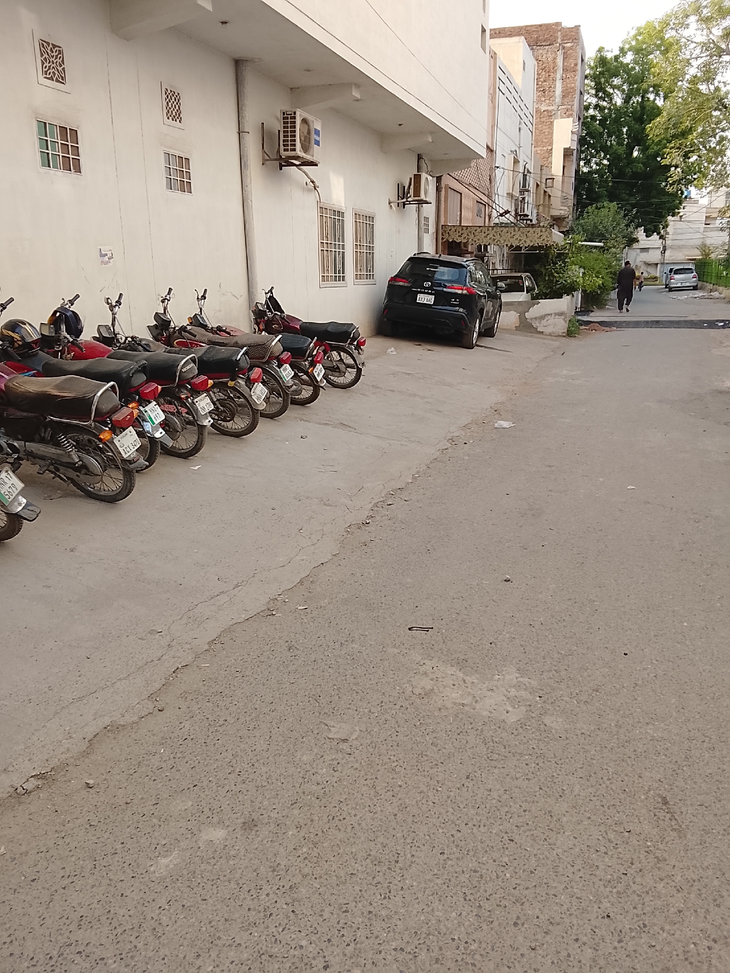 The scene is a street in Gulgasht Colony, Pakistan. A row of motorcycles is parked along the left side of the street, adjacent to a white building with windows and an air conditioning unit. A black car is parked behind the motorcycles, slightly angled into the street. A person is walking away from the camera on the right side of the street towards a white car parked further down the road. The street appears to be paved and shows signs of wear. The environment includes trees and buildings in the background.