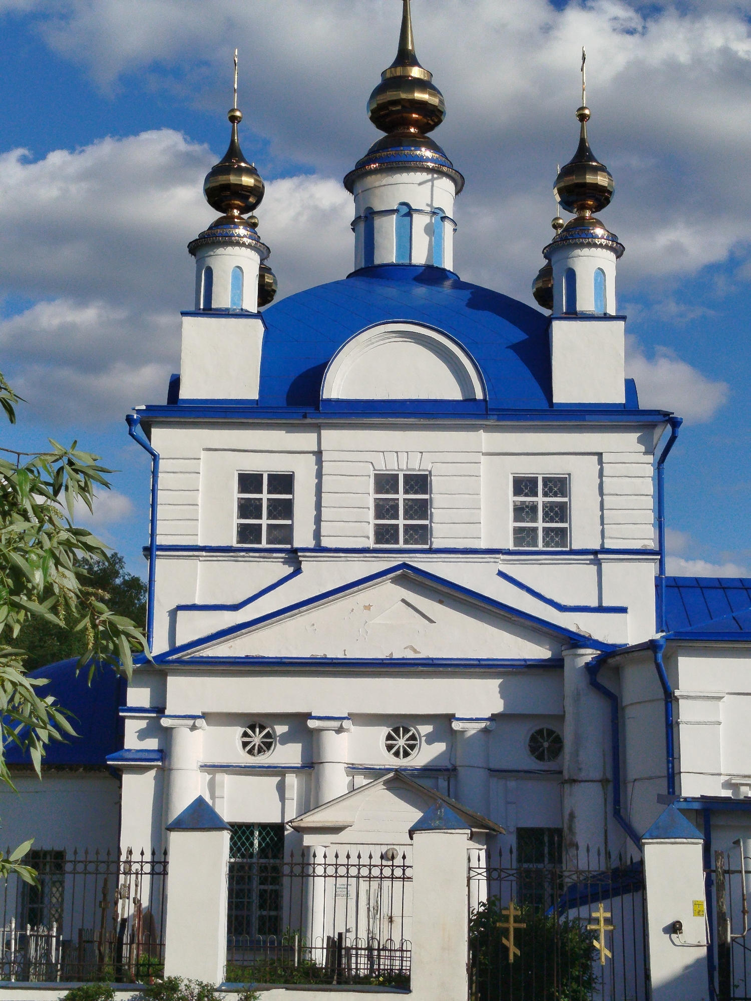 The image shows the Ascension Church in Mamontovo, Russia. The church features white walls with blue trim and a large blue dome topped with a smaller white structure and a golden onion dome with a cross. Two smaller towers flank the central dome, each similarly topped with a golden onion dome and cross. The building has rectangular windows with a grid pattern. The façade includes decorative circular windows and a triangular pediment over the entrance. A metal fence surrounds the church grounds, and a few plants and trees are visible in the foreground. The sky is blue with white clouds.