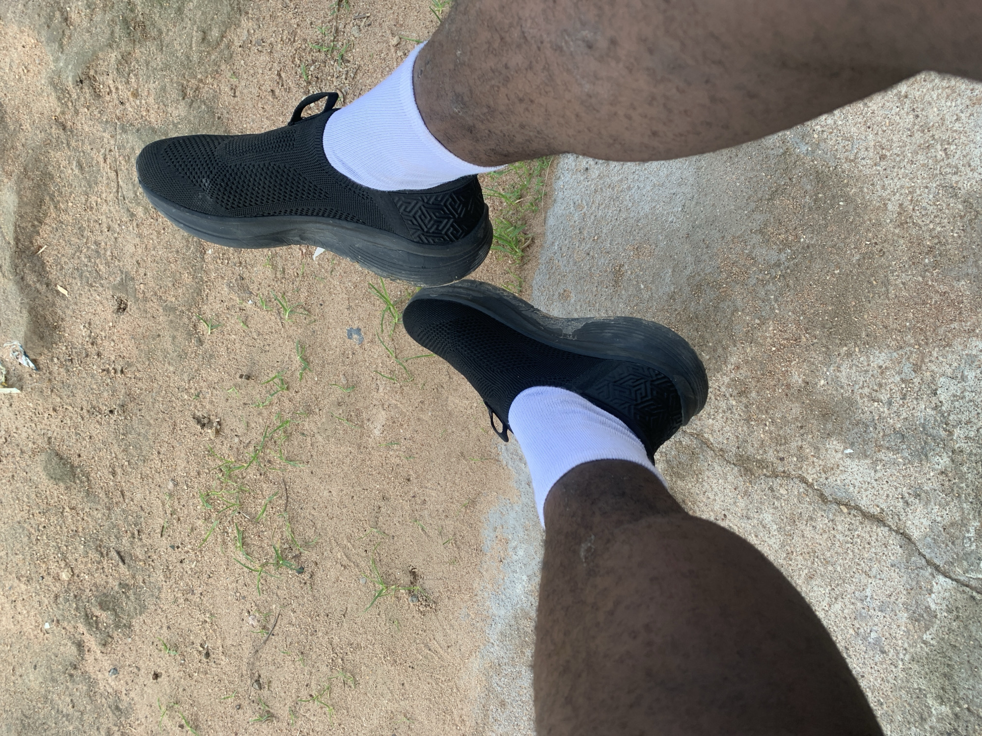 The image shows a person's legs and feet, wearing black slip-on athletic shoes and white ankle socks. The person is sitting on a concrete surface with a sandy area containing sparse green grass in the background. The lighting suggests it is daytime. The location is Jalingo, Nigeria.