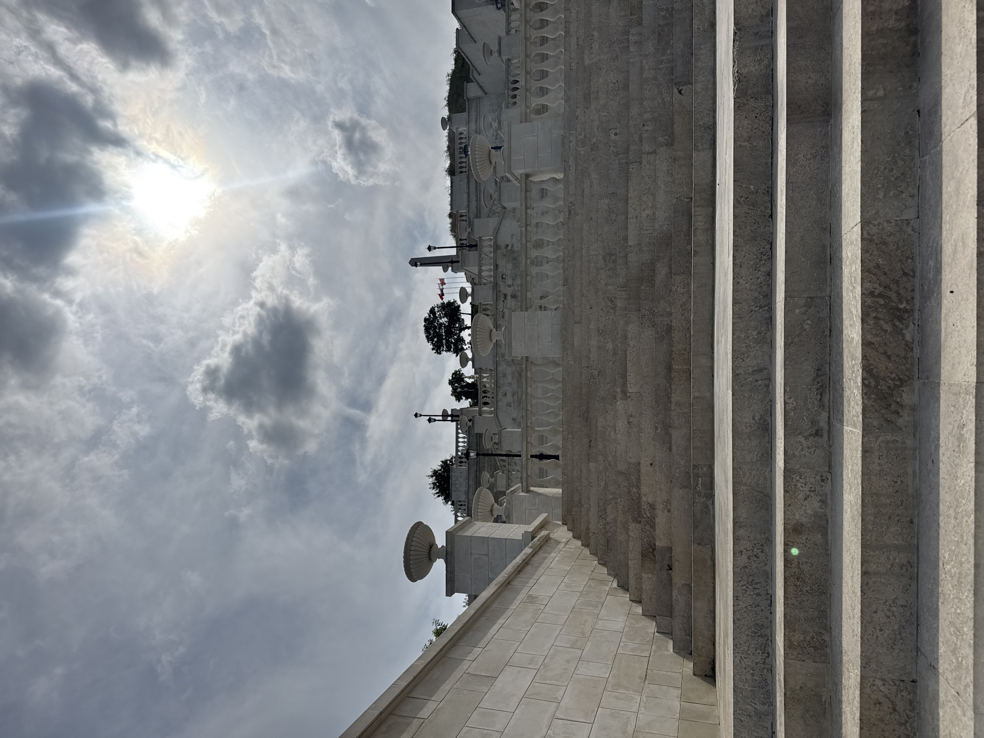 The image depicts a grand stone staircase leading upwards towards a tiered structure adorned with decorative urns and balustrades. The architecture suggests a historical or monument-like setting. Above, the sky is partially obscured by clouds, with the sun prominently shining through, creating a bright glare and lens flare. In the distance, silhouetted against the sky, are trees and flagpoles, one of which appears to be flying flags. The lower portion of the image is dominated by the textured stone steps. The scene is static, with no people visible. This is a view of the monumental stairway leading to the Mithridates Stairs in Kerch, Ukraine.