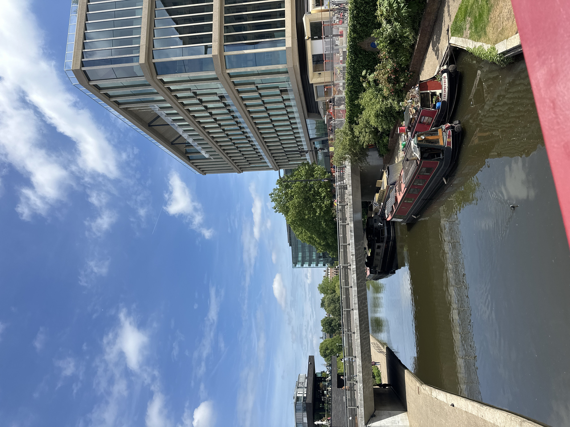 A slightly rotated photograph shows a canal in London, United Kingdom, with several barges moored along the bank. The barges are mostly red and black, with various items stored on their decks. The canal reflects the sky and the surrounding buildings.
Along the bank, there are trees and greenery. Adjacent to the canal, there is a pedestrian walkway with a metal railing. In the background, a large, modern office building with a grid-like facade dominates the scene. The sky is blue with scattered white clouds, with a faint contrail visible.
The photograph appears to be taken from a bridge or an elevated vantage point, with a red bridge railing partially visible in the upper right corner of the frame. Below the bridge, there is an entrance way on either side of the water.