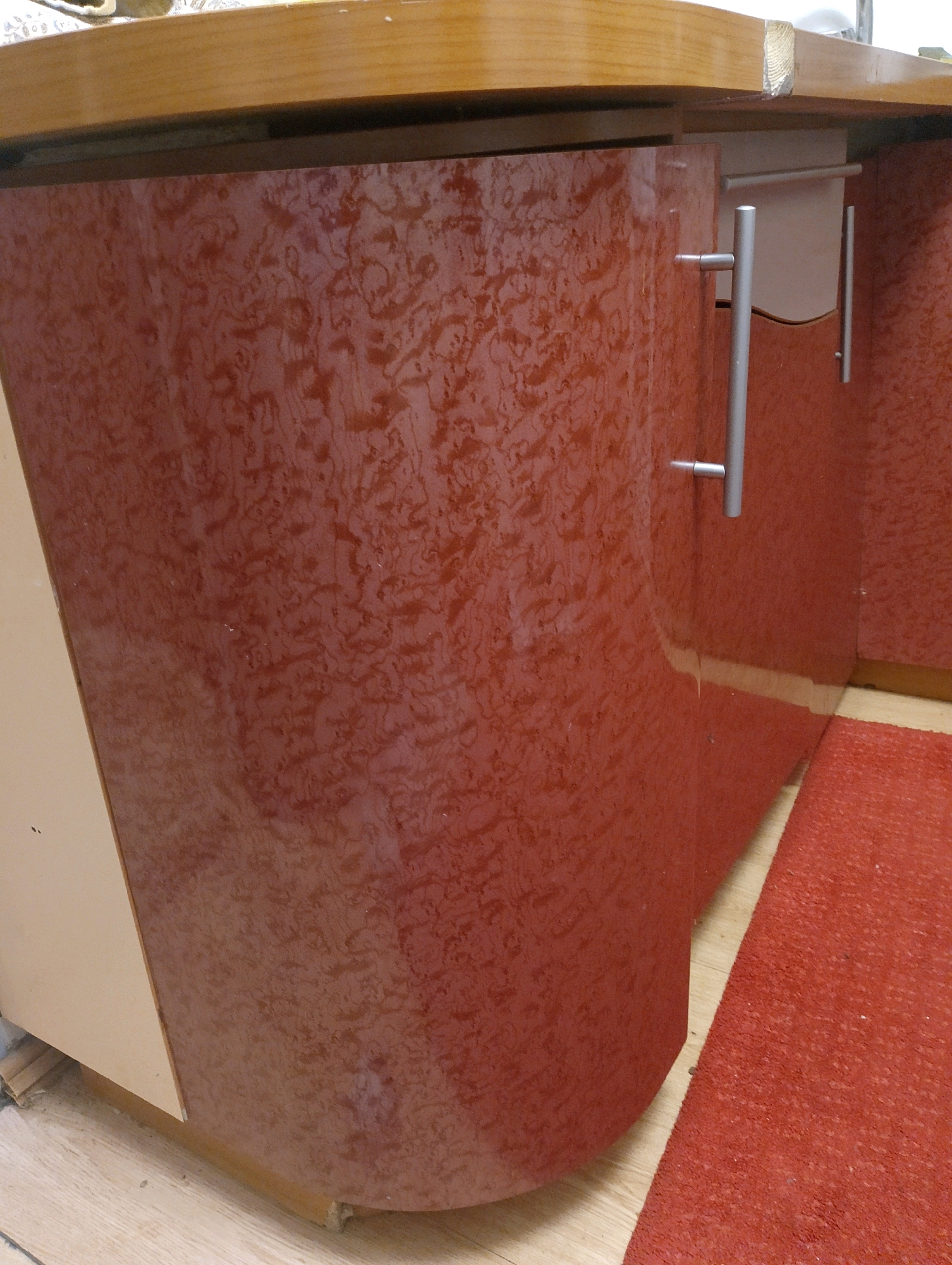 The image shows a close-up of a curved, reddish-brown cabinet with a marbled pattern. The cabinet has a light-colored edge on the left side, possibly a drawer or panel. Two silver handles are attached to the front of the cabinet, one larger and vertical, the other smaller and horizontal. The cabinet is situated on a light-colored wooden floor, and a red rug is visible in the lower right corner. A light-colored countertop is above the cabinet. The image does not provide any information about the city of Petrovskoye, Russia, or its inhabitants.