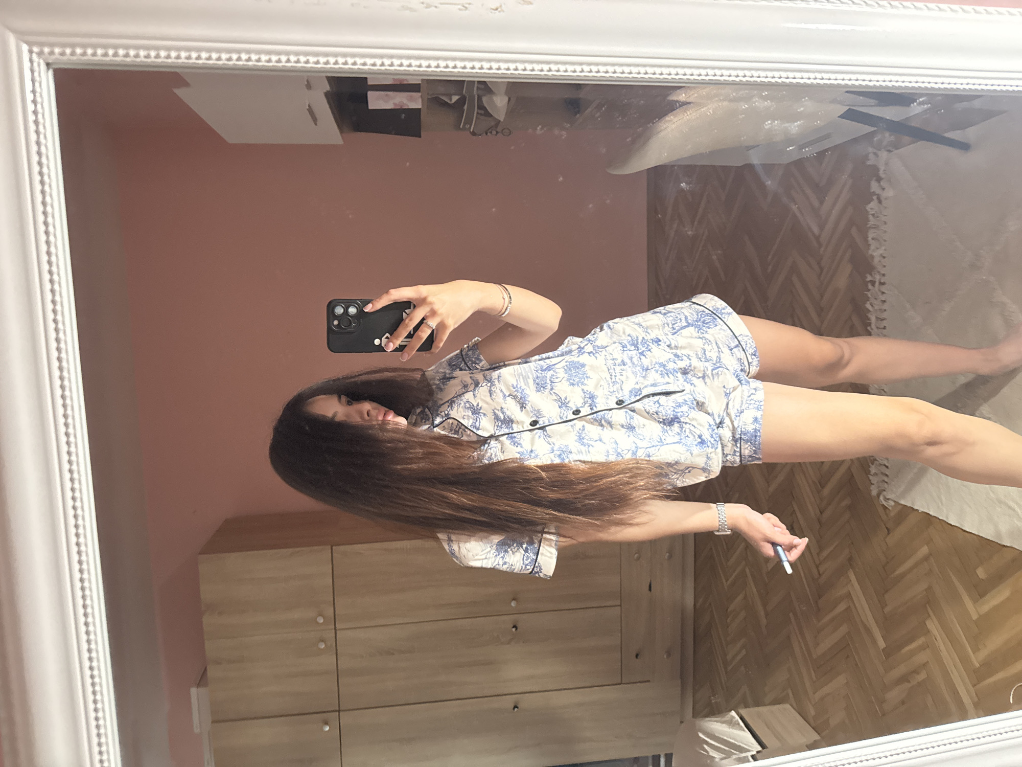 A young woman with long, dark brown hair is taking a selfie in a bedroom. She is wearing a white and blue patterned pajama set. Her left hand is holding a smartphone, and her right hand is holding a white object with a blue tip, possibly a pen or a cosmetic item. She is standing in front of a large white ornate mirror. In the reflection of the mirror, a wooden closet is visible to the left, and shelves with various items are on the right. The floor is made of wooden parquet. The walls are painted a soft pink. The image appears to be taken indoors, in a residential setting, likely in Plovdiv, Bulgaria, given the context of the request.