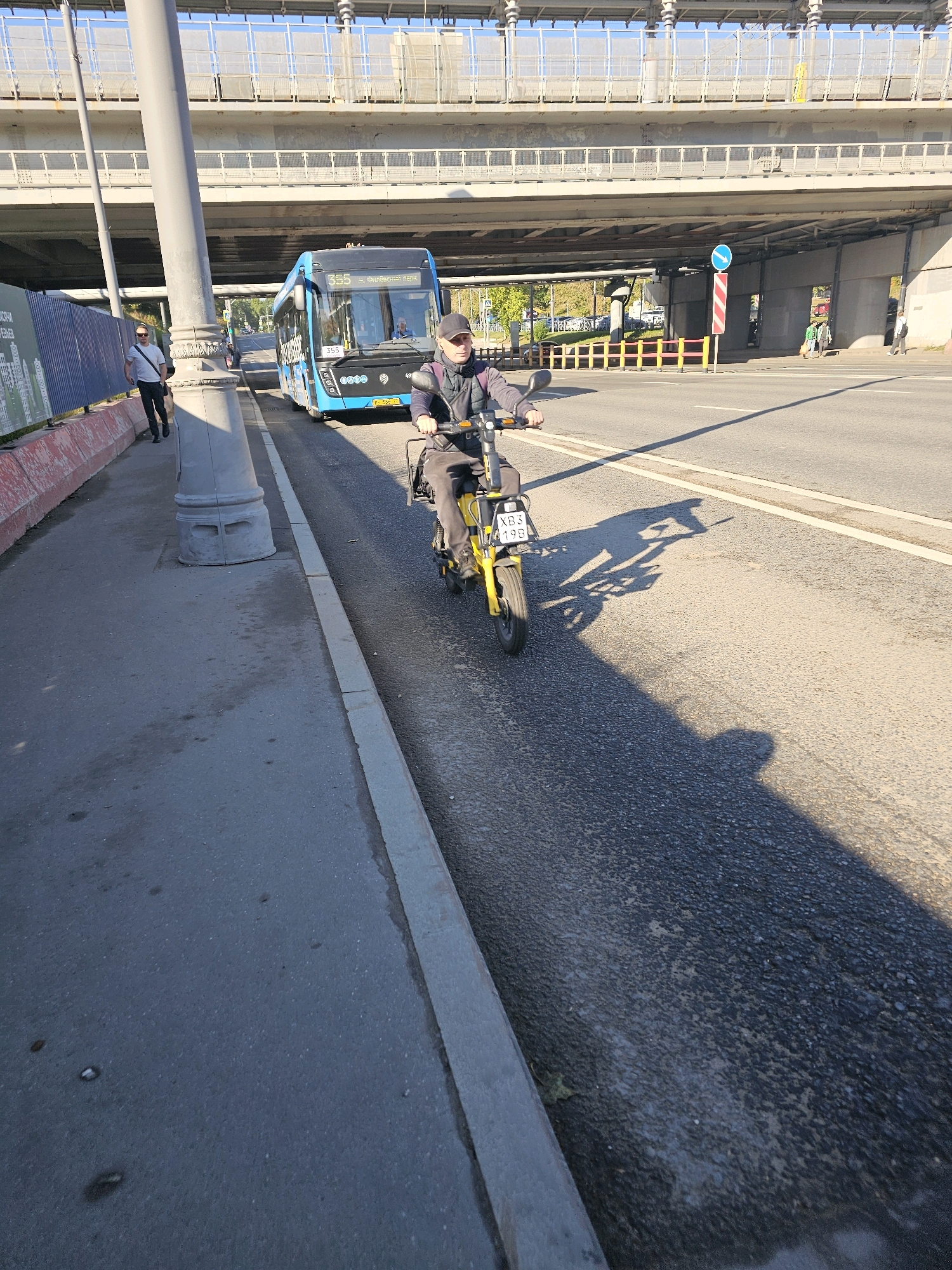 A man in a dark baseball cap and jacket rides a yellow electric scooter on a paved road. He is wearing dark pants and shoes. The scooter has a license plate that reads "XB3 19B". A blue bus with the number "355" is visible to the left of the scooter, driving in the same direction. The bus has "ФУНДАМЕНТАЛЬНАЯ" written on its side. In the background, there is an elevated highway structure with concrete supports. A sidewalk with a grey curb runs along the left side of the road, and a person in a white t-shirt and black pants is walking on it, carrying a dark bag over their shoulder. The sun is casting shadows on the road, indicating it is likely daytime. There is a blue directional sign with a white arrow pointing to the right on a pole near the highway overpass. This scene appears to be in Moscow, Russia, given the style of the bus and the visible text.