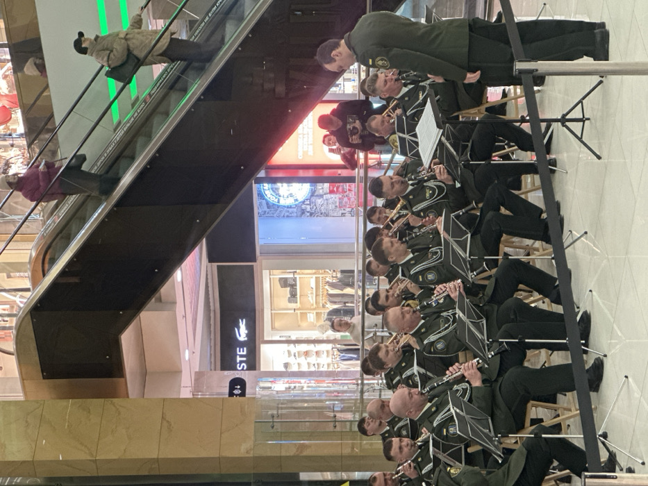 A military-style band performs indoors at a multi-story shopping mall in Kyiv, Ukraine. The band consists of approximately fifteen musicians, primarily brass and woodwind players, including clarinets and trombones. They are seated on wooden chairs with music stands in front of them, wearing dark green uniforms with insignia. A conductor, also in a dark green uniform, stands facing the band with his back to the viewer. A black rope barrier delineates the performance area. In the background, escalators ascend, transporting two individuals. Retail stores line the upper level, with a prominently visible "Lacoste" store sign. Several other individuals are visible in the background, some observing the band, including one person holding a camera. The environment features light-colored tiled floors, polished columns, and overhead lighting.
