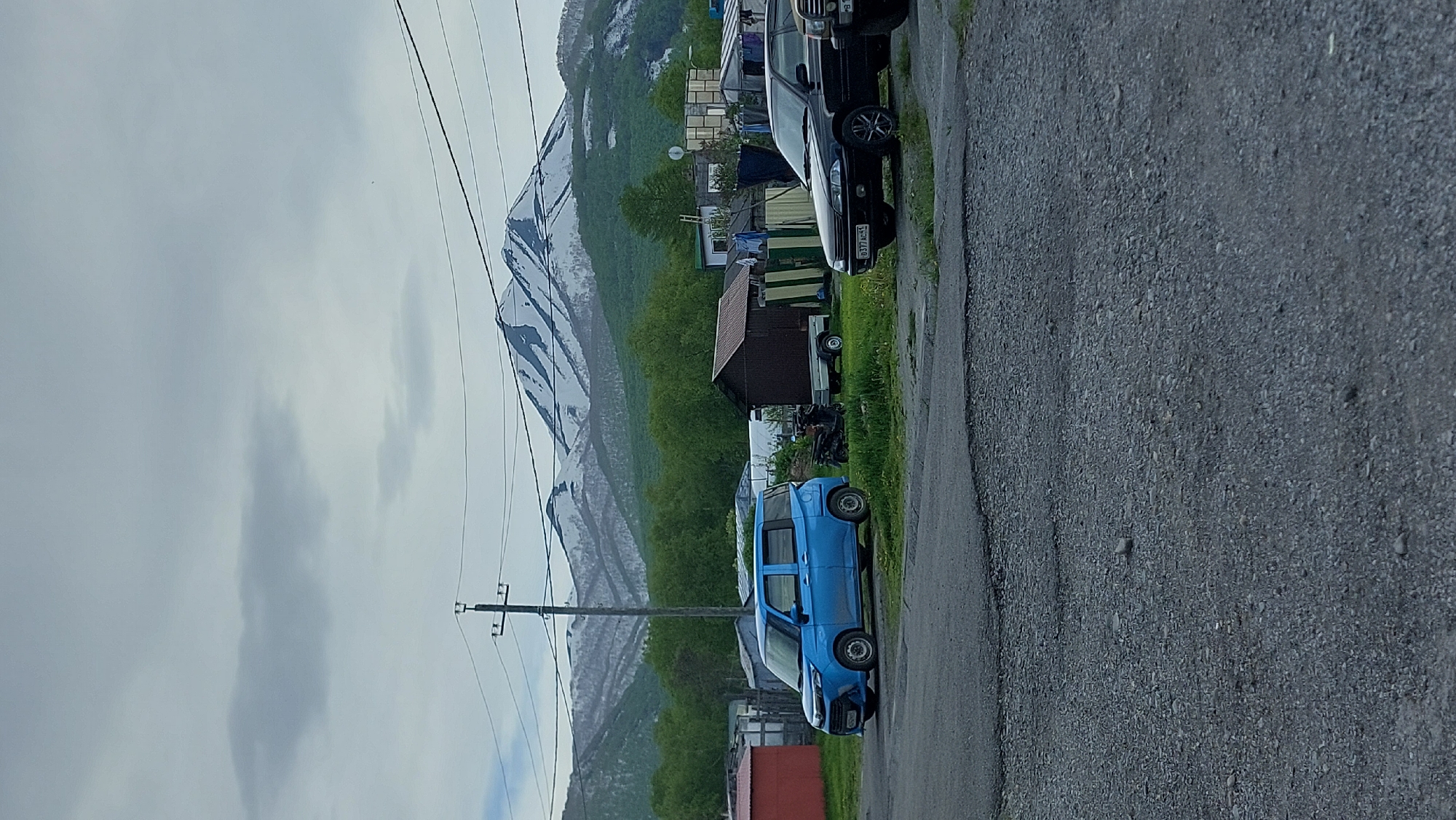 The image shows a rural scene in the city of Zeliony, Russia, with a large, snow-capped mountain in the background. In the foreground, there are several vehicles parked on a gravel road. A blue minivan is parked on the left, facing left. A black sedan is parked to the right of the minivan, facing left. A small utility trailer is attached to the back of a dark-colored vehicle partially visible on the far right. There are also several buildings and structures, including what appears to be a small shed and a larger building with a corrugated metal roof. The overall impression is one of a remote, somewhat rustic environment.