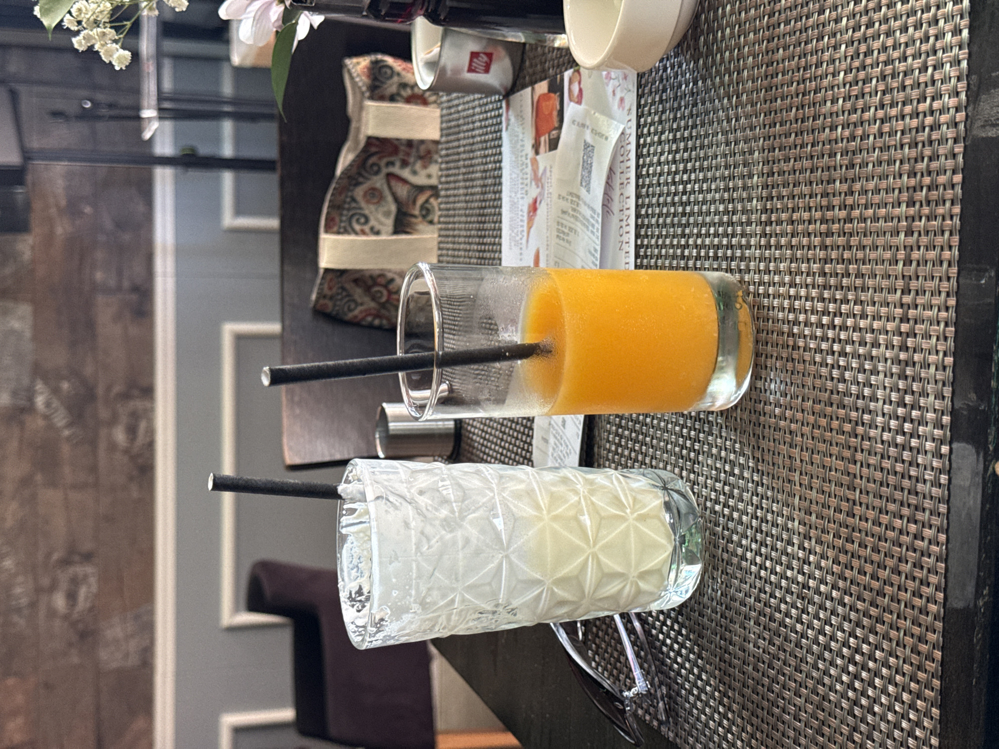 A close-up, eye-level shot shows two glasses of drinks on a woven placemat. The glass on the left contains a frothy, pale yellow beverage, possibly a smoothie or milkshake, with a black straw. The glass on the right holds a vibrant orange juice, also with a black straw. A pair of sunglasses rests on the left side of the placemat. In the background, a patterned bag with a cat's face motif is visible on a chair, along with a small vase of white flowers and a dark bottle. The setting appears to be a cafe or restaurant. The image provides no specific geographical markers to pinpoint the location in Budva, Montenegro.