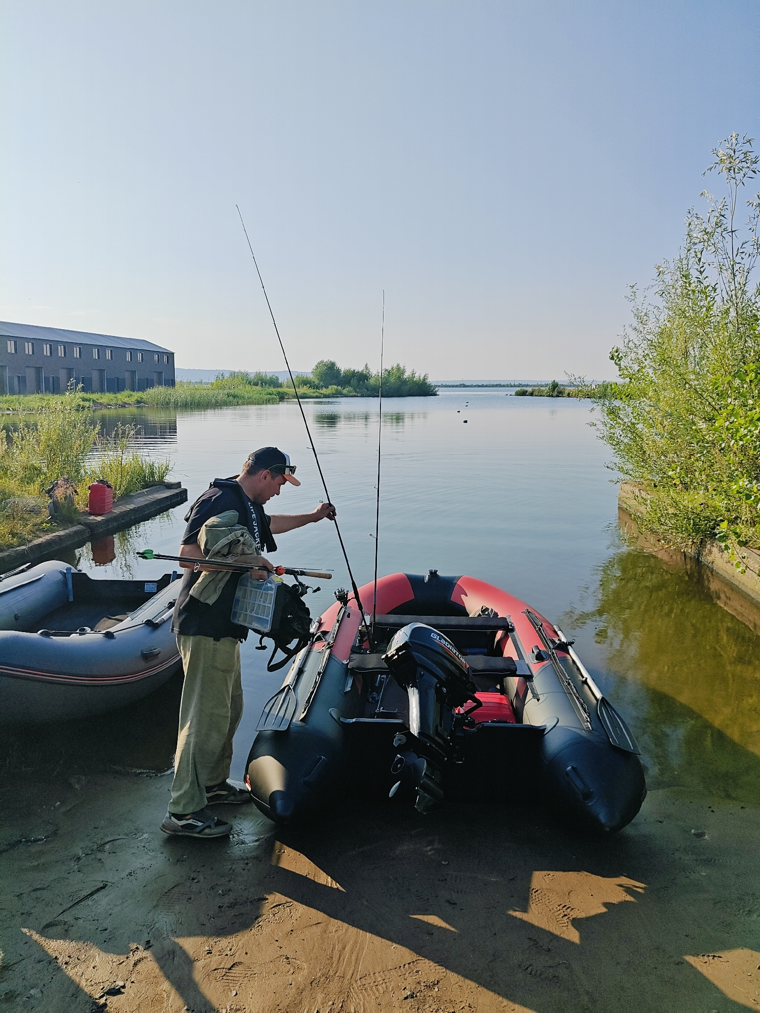A man is preparing fishing equipment by an inflatable boat at the edge of a body of water, likely a river or reservoir, in Chaikovsky, Russia. The man, wearing a black t-shirt, tan pants, a black baseball cap, and athletic shoes, is holding a fishing rod and a tackle box. He is standing next to a red and black inflatable boat equipped with an outboard motor labeled "GLADIATOR". Another inflatable boat, grey and black, is positioned behind him. The scene is set in a natural environment with green vegetation along the bank and a calm body of water reflecting the clear sky. In the background, a long, multi-story industrial building is visible. The ground is sandy and wet, indicating proximity to the water. The overall lighting suggests it is daytime, likely morning or late afternoon.