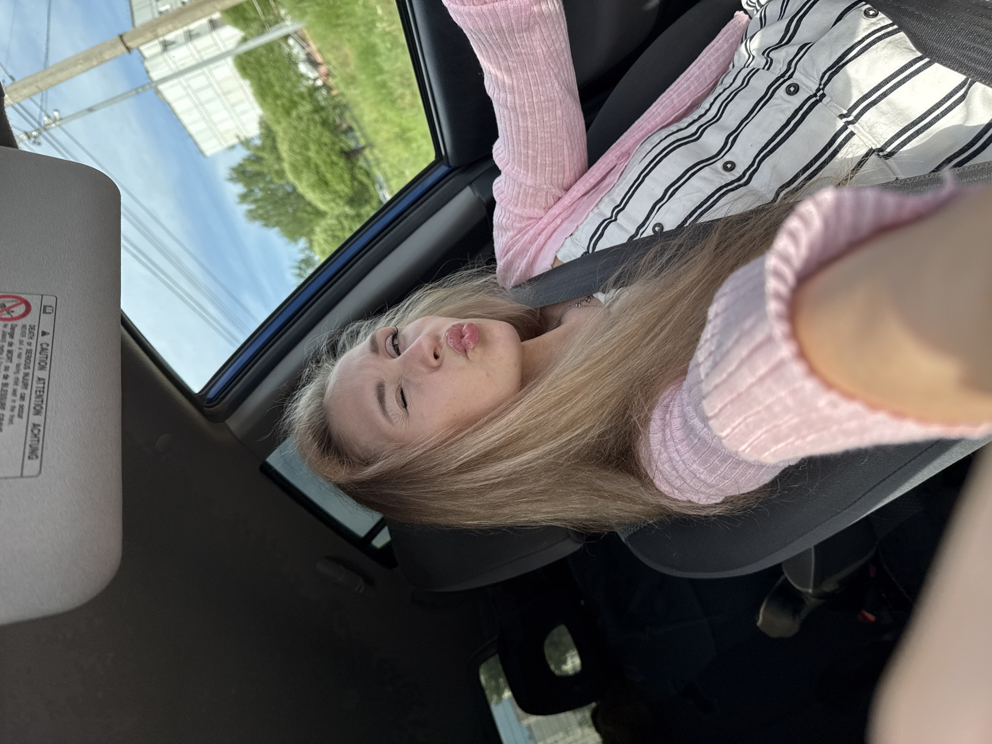 A blonde woman is taking a selfie in a car, likely in Chaikovsky, Russia, based on the OCR text. She is in the driver's seat, wearing a pink long-sleeved shirt over a striped button-down shirt. She is making a kissing face toward the camera. The car's sunroof is open, offering a view of buildings and trees outside. The car's sun visor and seatbelt are visible.