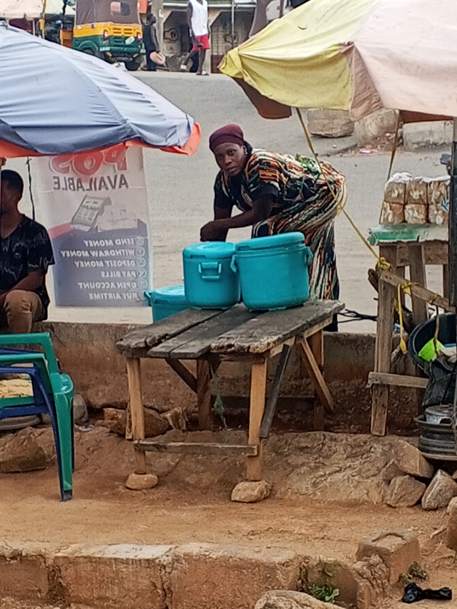 The scene depicts a woman standing behind a table with three blue food containers in Gwong, Nigeria. She is dressed in a patterned dress and a maroon headwrap. Behind her to the right, there is a table with stacks of bread. A faded yellow and white umbrella provides shade. To her left, a man sits near a poster that reads "POS AVAILABLE" along with reversed text related to money transactions. Another umbrella is visible on the left of the poster. A commercial vehicle is visible in the background, and the ground is uneven, with exposed rock and dirt.