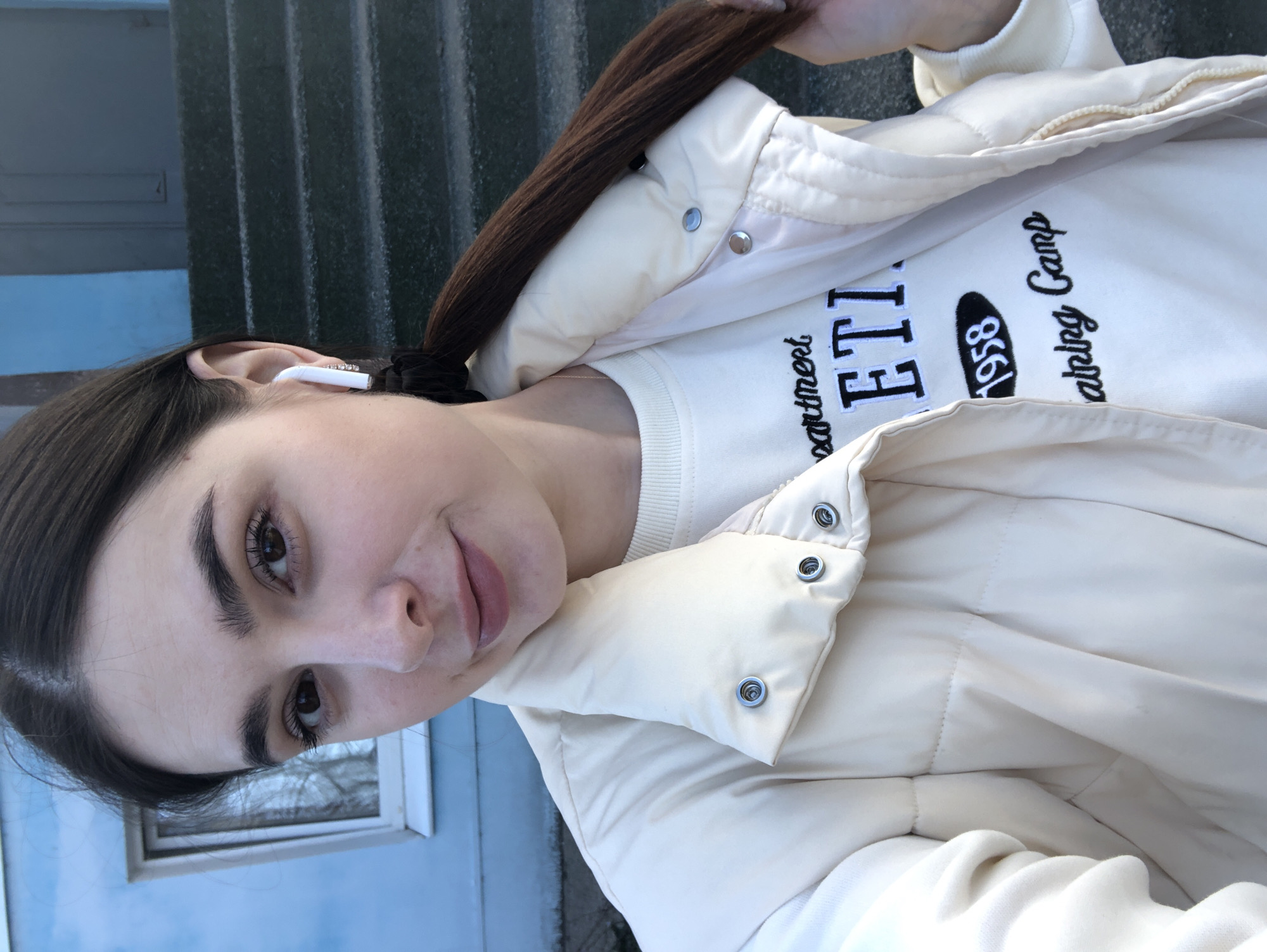 A young woman with dark hair, partially pulled back into a ponytail, is taking a selfie outside. She is wearing a white sweatshirt with black lettering that includes "1958" and a light-colored, quilted jacket. An earbud is visible in her left ear. The background features a blue building with a small window and grey concrete steps. The location is likely Chaikovsky, Russia.