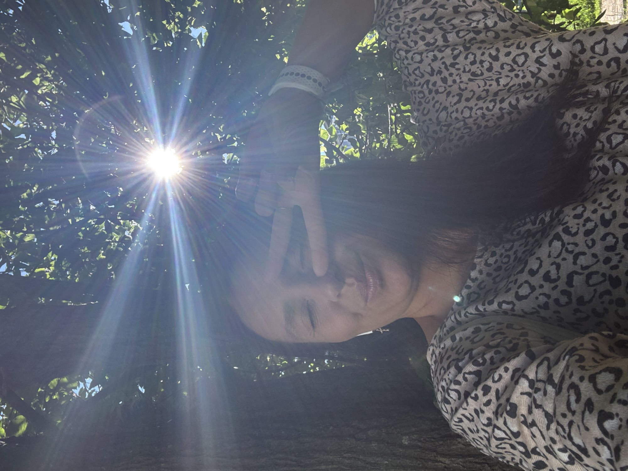 A medium shot captures a woman with long dark hair, wearing a leopard-print top and a white beaded bracelet on her right wrist, as she poses for a selfie outdoors. She is positioned under the dappled sunlight filtering through the leaves of a large tree. Her eyes are closed, and she holds her right hand up to her face in a peace sign gesture, her fingers obscuring part of her forehead. Her lips are pursed, giving her a playful expression. The bright sun is prominently featured, creating a starburst effect with lens flares that spread across the image. The background shows the textured bark of a tree to the left, dense green foliage, and a hint of blue sky. The overall setting appears to be a natural outdoor environment, possibly a park or a backyard, within the city of Chaikovsky, Russia, given the context provided.