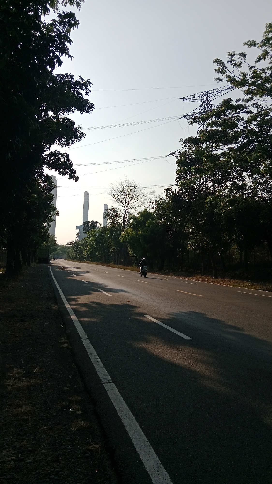 The image captures a road scene in Bhinar, Indonesia, on a clear day with bright sunlight casting shadows. A paved road with a white line marking the edge runs diagonally from the bottom left to the upper right of the frame. On the right side of the road, a dark green motorcycle is being ridden by an individual wearing a helmet and dark clothing. Trees line the right side of the road, and beyond them, a large metal electricity pylon is visible. In the distance, on the left side of the road, two tall, slender white structures resembling smokestacks from a power plant are prominent. Trees also line the left side of the road, obscuring the lower portions of these distant structures. The sky is bright and clear, with power lines running across it. The overall impression is one of a semi-urban or industrial area with significant vegetation along the roadside.
