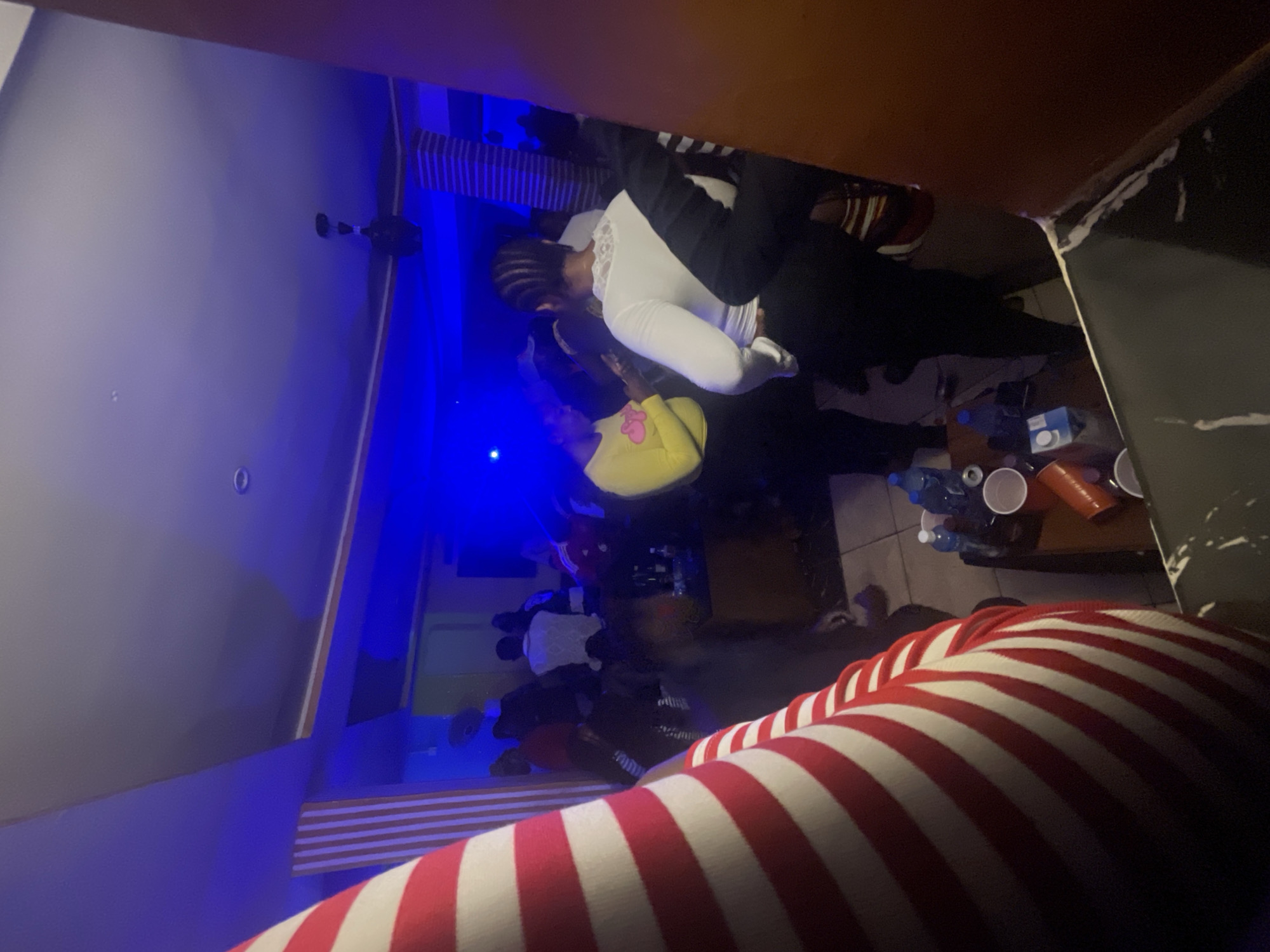 The image shows a group of people in what appears to be a nightclub or bar in Durumi, Nigeria. The room is dimly lit, with prominent blue and purple lighting. A ceiling fan is visible in the upper portion of the image. In the foreground, a person wearing a red and white striped garment is partially visible, facing away from the camera.
In the mid-ground, several individuals are gathered. One woman on the left is wearing a yellow top and black pants. Another woman in the center is wearing a white top with lace trim and dark pants. Her hair is styled in braids. Other individuals are seen in the background, some of whom appear to be standing or sitting.
To the right of the central figures, a table or counter is visible with several plastic cups, bottles of water, and what may be alcoholic beverages. The overall atmosphere suggests a social gathering or party. The environment appears to be an indoor commercial space, possibly a lounge or entertainment venue. The image quality is somewhat grainy, likely due to low light conditions.