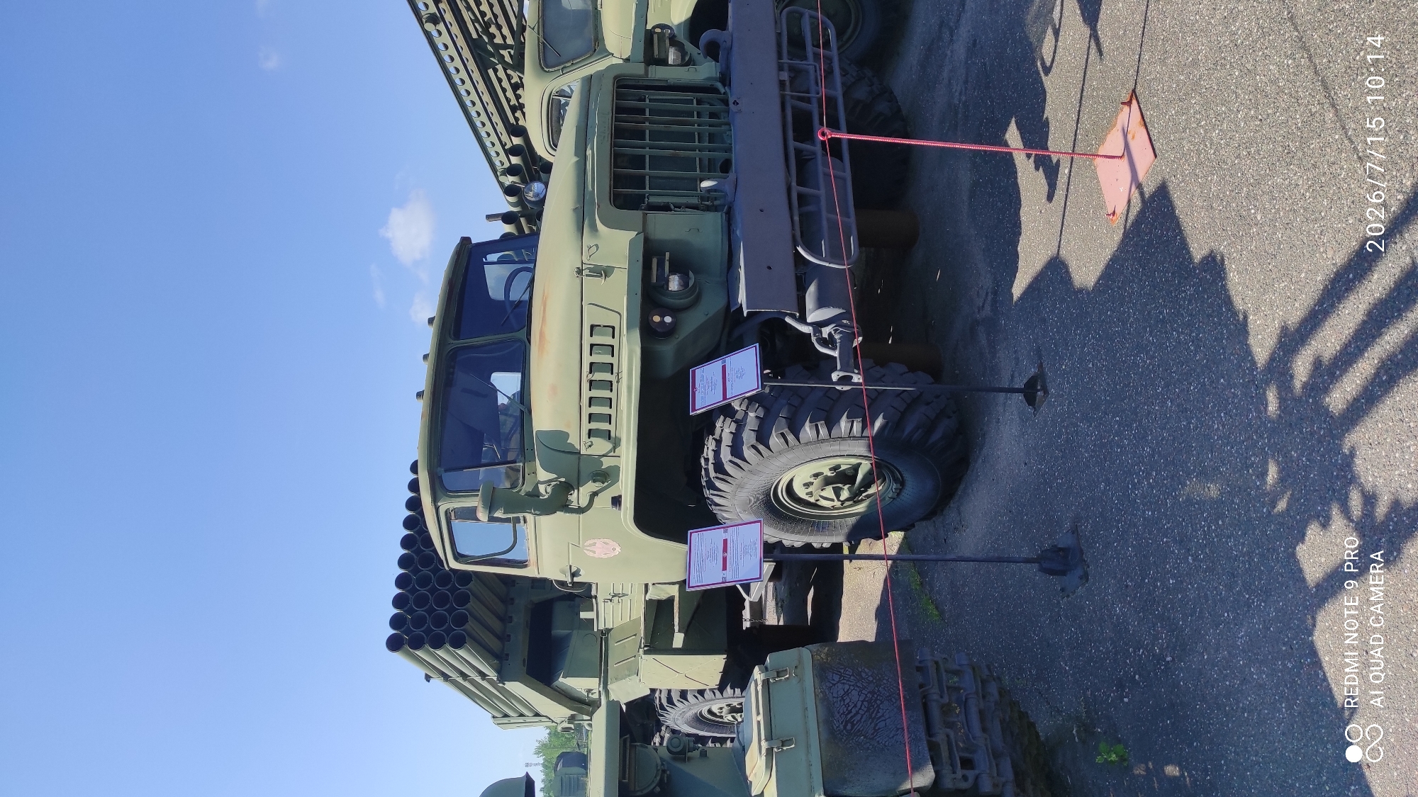 A military rocket launcher truck is parked on an asphalt surface under a clear blue sky. The truck is painted in a camouflage green color and has a large rack of rockets mounted on its rear. Two signs with text in a foreign language are placed in front of the truck's tires. The truck is part of an outdoor exhibition in the city of Лашанскі сельскі Савет, Belarus. The lighting suggests it is daytime, with shadows cast by the truck and signs onto the ground.