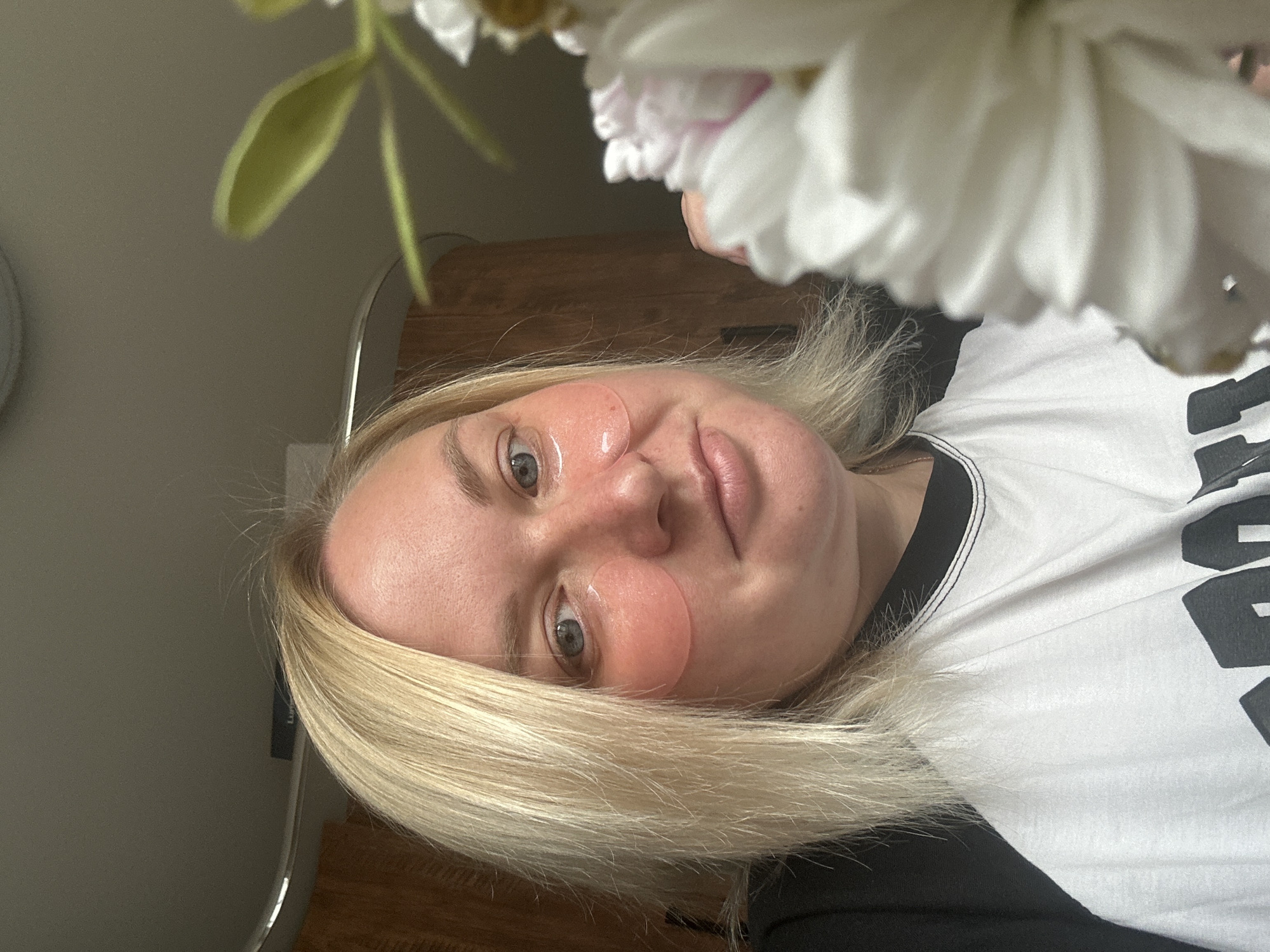 A blonde woman with blue eyes is shown in a close-up, selfie-style shot. She is wearing pink under-eye patches and a white t-shirt with black trim and lettering. Her hair is parted to the side and falls to her shoulders. The background appears to be an indoor setting with wooden cabinetry and a ceiling. A blurred white flower is visible in the foreground on the right side of the image. The location is not specified as Chaikovsky, Russia.