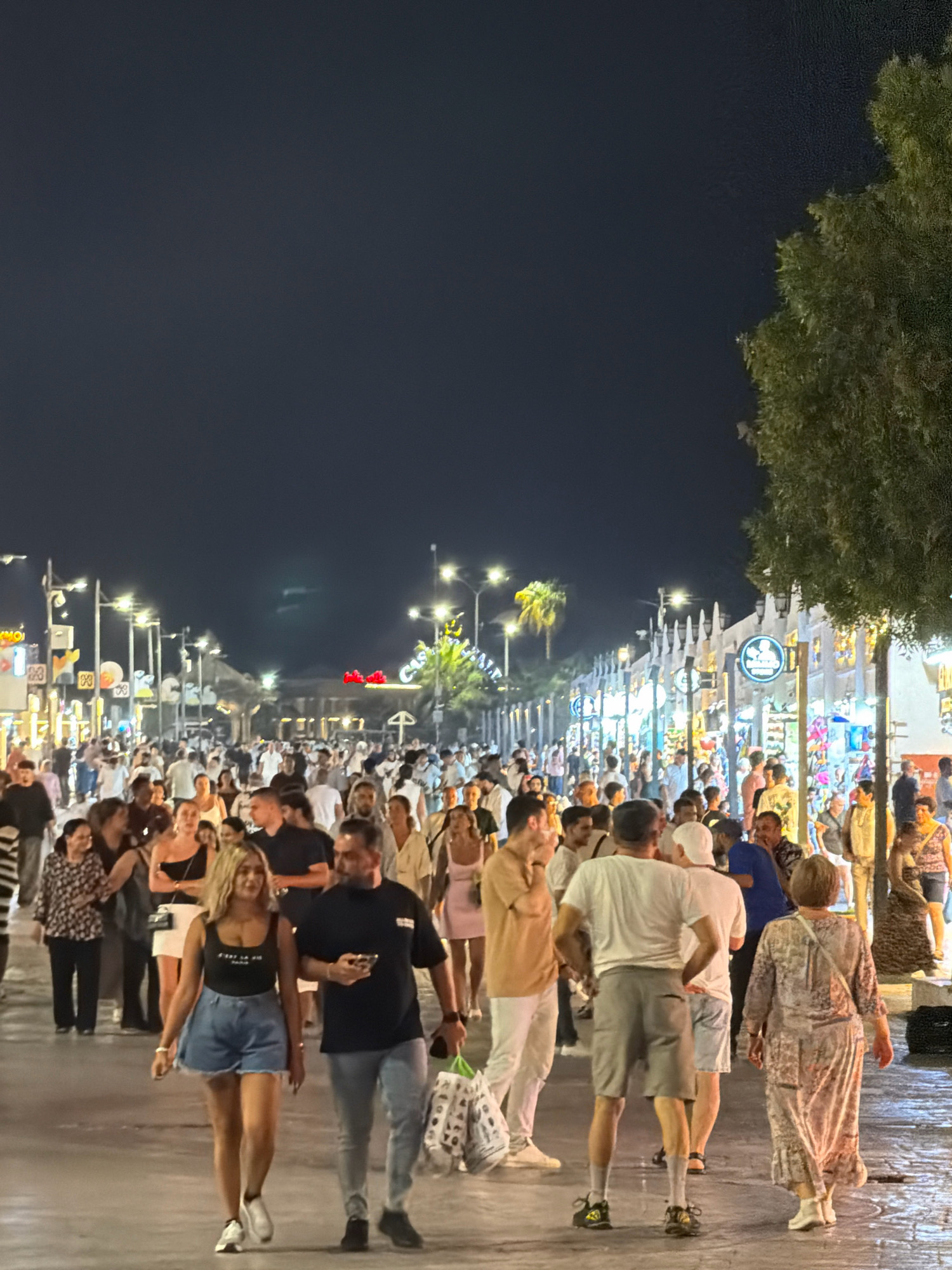 This image captures a bustling nighttime scene on a street in Sharm El Sheikh, Egypt, likely in the Naama Bay area, known for its lively atmosphere. A dense crowd of people, a mix of locals and tourists, are seen walking along a paved pathway lined with shops and illuminated by streetlights and colorful signs. The architecture visible suggests a resort town setting, with buildings featuring white facades and decorative elements. Palm trees are scattered along the street, adding to the tropical ambiance. Many individuals are casually dressed, reflecting the warm climate. The overall impression is one of vibrant activity and social gathering under the night sky.
