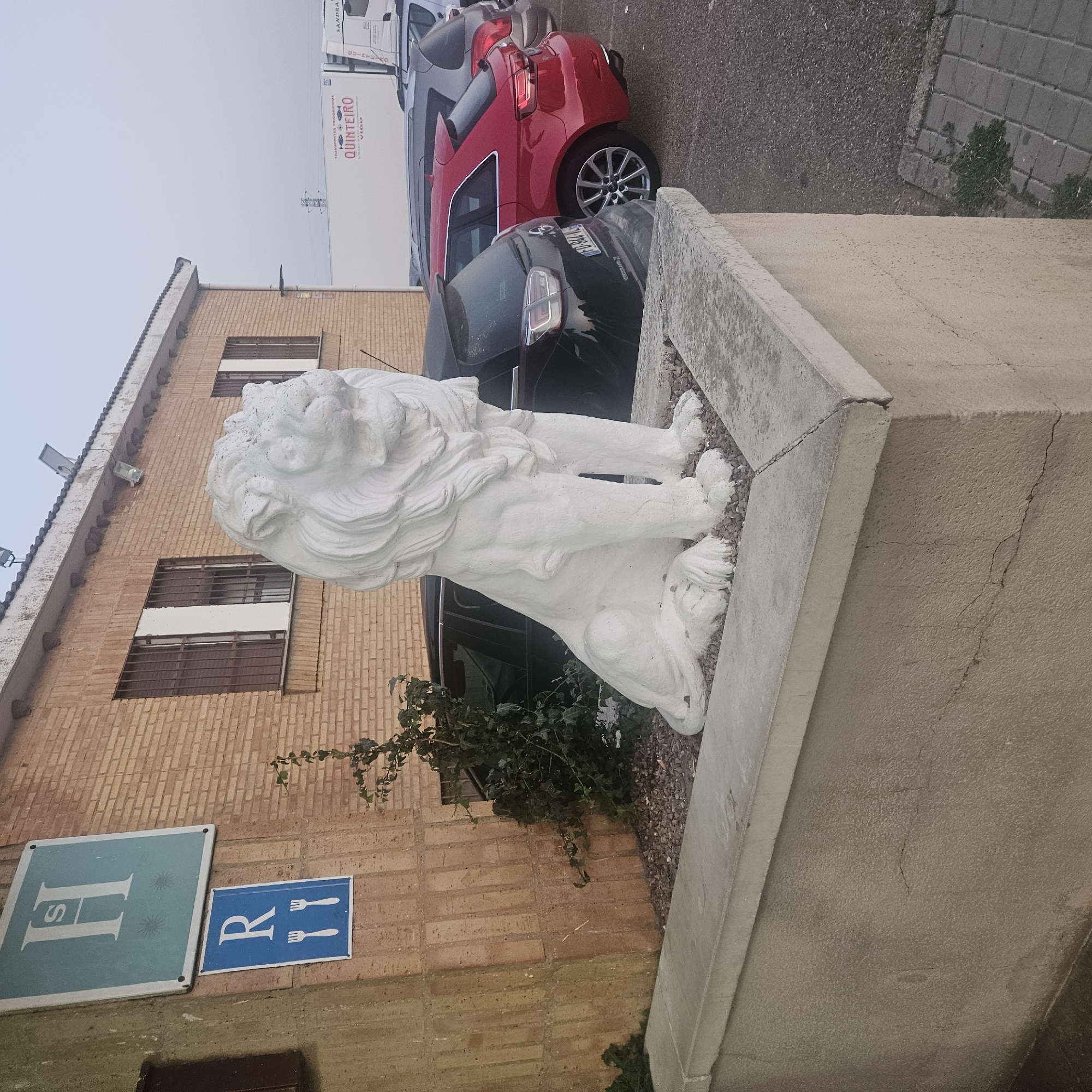The image depicts an outdoor scene in Becilla de Valderaduey, Spain, featuring a white lion statue in the foreground. The statue is seated on a grey concrete platform covered with small, dark pebbles. Behind the statue, a brick building with windows is visible. On the side of the building, two signs are mounted: a larger, light blue sign with a white "H" and two starbursts, and below it, a smaller blue sign with a white "R" and two forks. To the right of the building, parked cars are visible, including a red hatchback and a darker SUV. In the background, a white truck with "QUINTEIRO VIGO" printed on its side and a trailer is parked. The environment appears to be an overcast day.