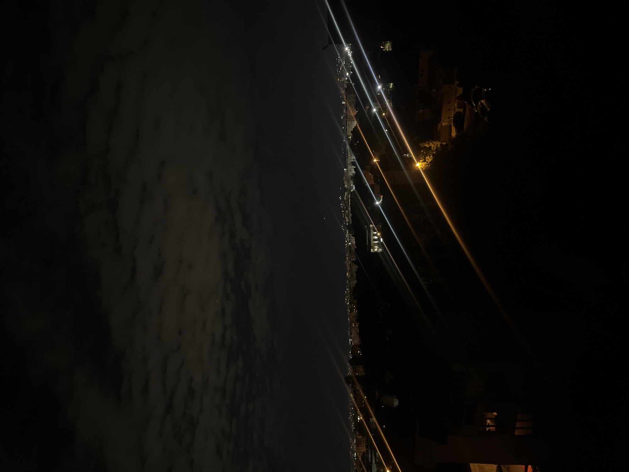 The image depicts a nighttime view of what appears to be a cityscape along a coastline. The left side of the image shows a cloudy sky, while the center features a line of lights suggesting a road or city streets. Bright light sources are visible, possibly streetlights or building lights. The right side is dominated by darkness, with some indistinct structures. Based on the presence of a city along what appears to be a coastline, the image is unlikely to be in Sofia, Bulgaria. The city is not coastal.