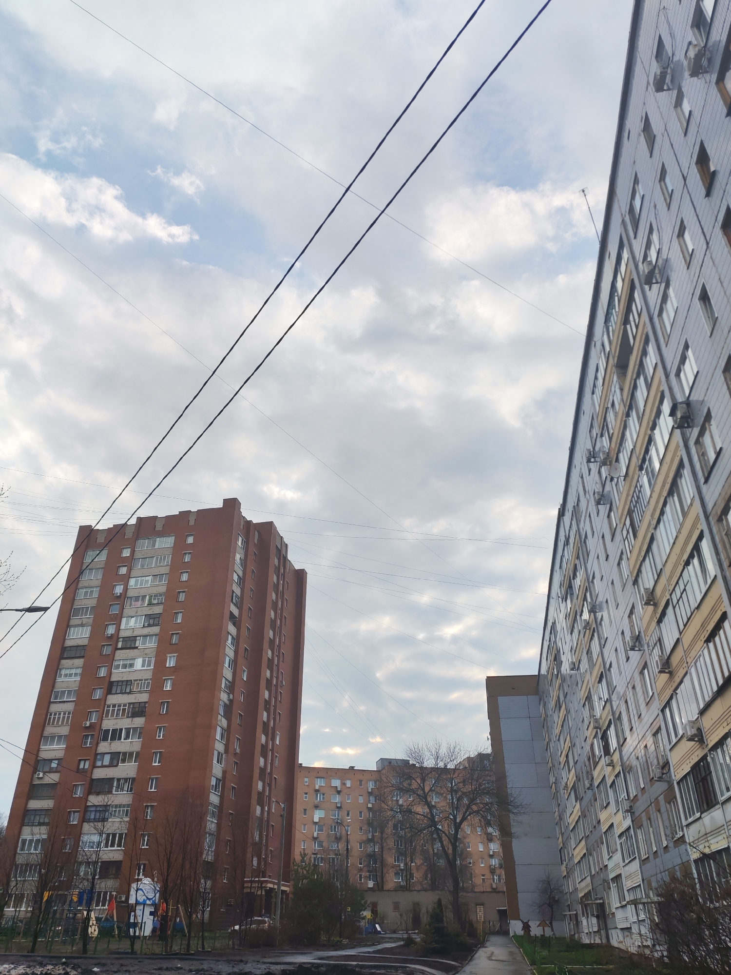 The image shows an urban scene in Izhevsk, Russia, dominated by two tall residential buildings under a cloudy sky with power lines overhead. The building on the left is a reddish-brown brick high-rise, while the one on the right is taller and has a gray facade with beige window frames and numerous air conditioning units. A playground is visible at the base of the brick building on the left, and there are trees lining the street between the buildings. The street appears wet, suggesting recent rain. Another residential building is seen further down the street between the two larger buildings. There are bare trees indicating it is likely autumn or early spring. No people are clearly visible in the frame.