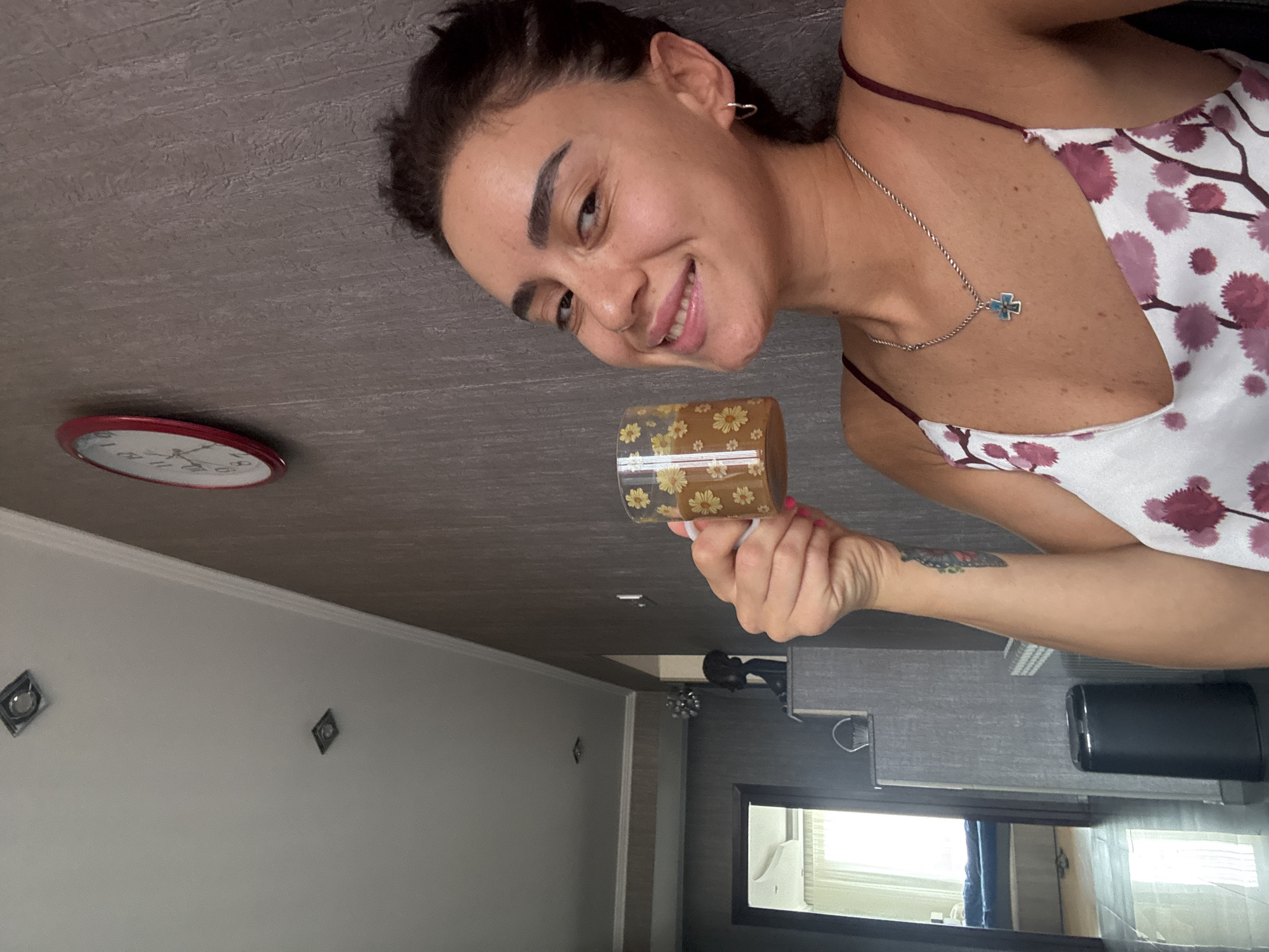 A woman is taking a selfie indoors, holding a patterned mug of coffee. She is smiling and looking towards the camera. The background shows a wall with a clock and some recessed lighting in the ceiling. In the distance, a doorway leads to another room. The woman is wearing a floral print top and a silver necklace. The lighting suggests it is daytime.