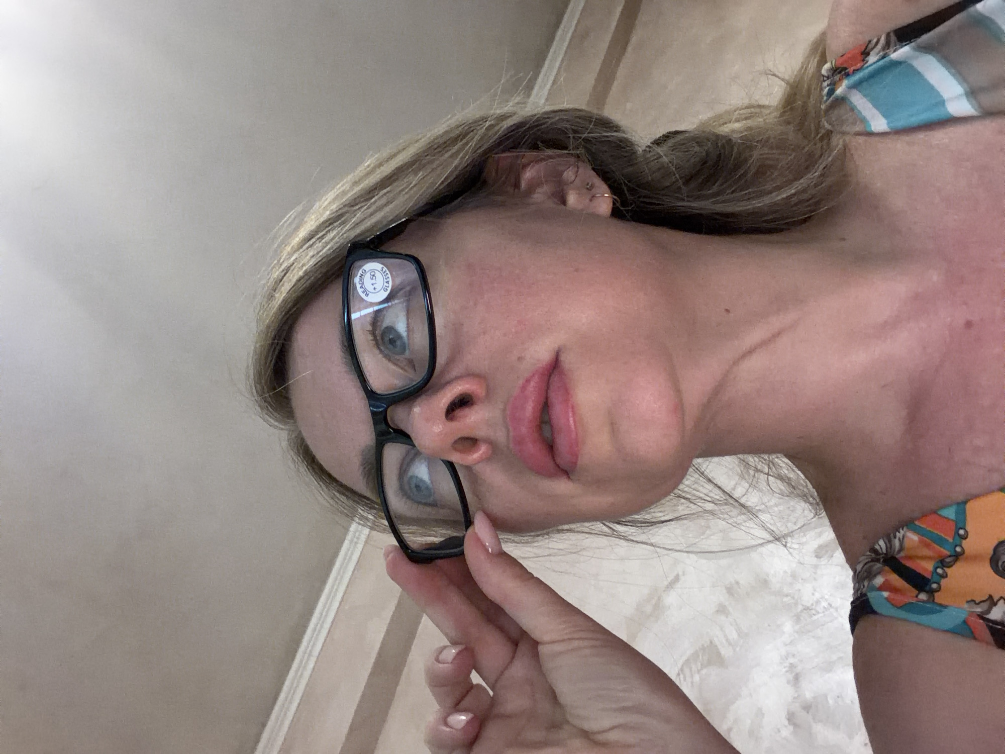 A medium shot shows a woman wearing black-rimmed reading glasses, adjusted downwards on her nose by her right hand. She has blonde hair and is looking upwards to the left. The glasses have a circular sticker on the left lens that reads "READING +1.50 GLASSES". She is wearing a colorful top with orange, blue, and black patterns. The background appears to be an indoor ceiling with some light fixtures. There is no indication of the city of Rome, Italy in the image.