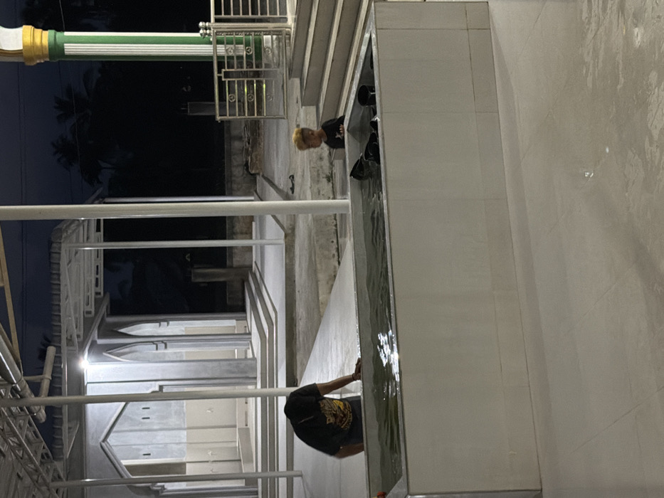 This image captures an illuminated ablution area within a mosque complex in the city of Bale, Indonesia, during nighttime. The scene is dominated by a rectangular, white-tiled water basin centrally located, containing water and several black ladles or scoops. An adult, visible from the torso up wearing a dark t-shirt, is leaning over the left side of this basin, with hands submerged or resting near the water's surface. To the right of the basin, a young child with distinctly blonde or light-colored hair and a dark t-shirt stands facing towards the basin or the adult.
In the background, a concrete structure featuring multiple arched openings and faucets, indicative of a wudu (ablution) station, is visible. This structure appears new or recently renovated, with clean lines and light gray finishes. White metal support columns and an overhead canopy extend across the scene. To the far right, a prominent architectural column with green and gold vertical stripes stands next to metal railings and a set of concrete steps. The ground is paved with light-colored tiles, exhibiting wet patches and reflections from the artificial lighting. Beyond the constructed area, the dark night sky and silhouettes of distant trees are discernible.