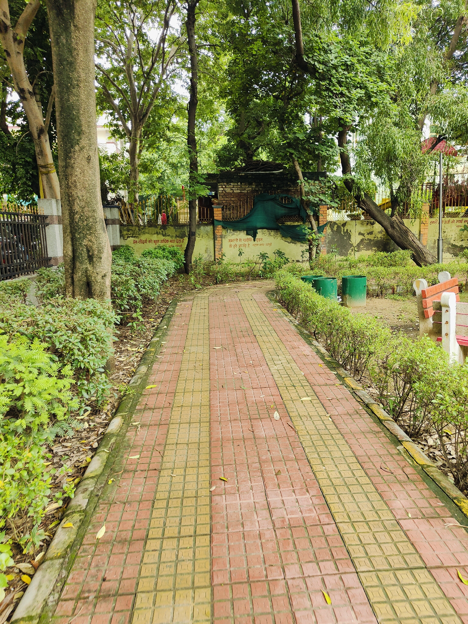 The image depicts a paved walkway in a park setting in Gurgaon, India. The walkway is made of square tiles in alternating colors of red and yellow, with a yellow border along its edge. Flanking the walkway are trimmed green bushes. In the background, there are several trees with lush green foliage. Behind the bushes on the right side of the walkway, a park bench with orange and white elements is visible, as are two green cylindrical trash receptacles. A brick wall runs along the background, partially obscured by greenery and a dark green tarp. The wall has some writing on it, which appears to be in Hindi. The overall environment suggests a public park or garden area, likely maintained for recreational use, located in an urban or suburban area of Gurgaon.