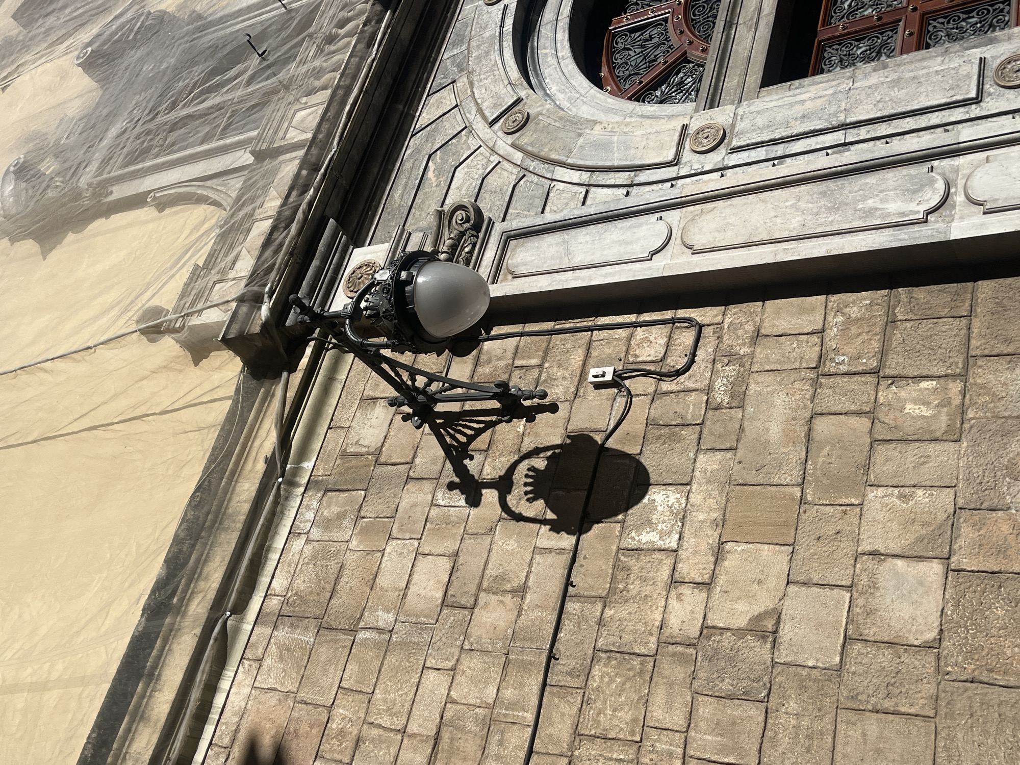 The image depicts a stone building facade in Barcelona, Spain, partially obscured by scaffolding covered in a beige tarp. A vintage-style iron lamppost is mounted on the wall, casting a shadow on the stonework below. A window with ornate metal grillwork is visible above the lamppost. The facade is constructed of rectangular stone blocks, and the overall scene suggests an urban environment undergoing construction or renovation.