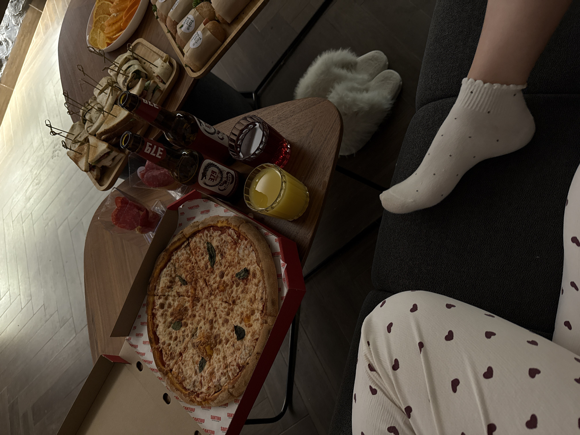 This indoor shot, set in Yerevan, Armenia, depicts a casual meal or relaxing evening. A wooden table holds a spread of food and drinks. In the foreground, a large, cheesy pizza, garnished with basil leaves, rests in an open cardboard box with "SANTINO" printed on the inner lining. Nearby, slices of cold cuts are visible.
Two bottles, one clearly labeled "GLE" and "CHERRY," and another also with "GLE," are positioned next to two glasses containing a red beverage and a yellow drink, possibly juice. Further back on the table, several small sandwiches or wraps, secured with toothpicks, are arranged on a wooden serving tray. Another white bowl holds slices of oranges or other citrus fruit. Packaging elements visible in the background include the words "Take" and "Eat."
On the right side of the frame, a person is seen relaxing on a dark gray sofa. Their left leg is visible, clad in a white ankle sock with small dark polka dots and a ruffled top. Their lower body is covered by white pajama bottoms featuring a pattern of small dark hearts. A pair of fluffy white slippers can be spotted on the floor near the sofa. The scene conveys a sense of comfort and a leisurely evening.