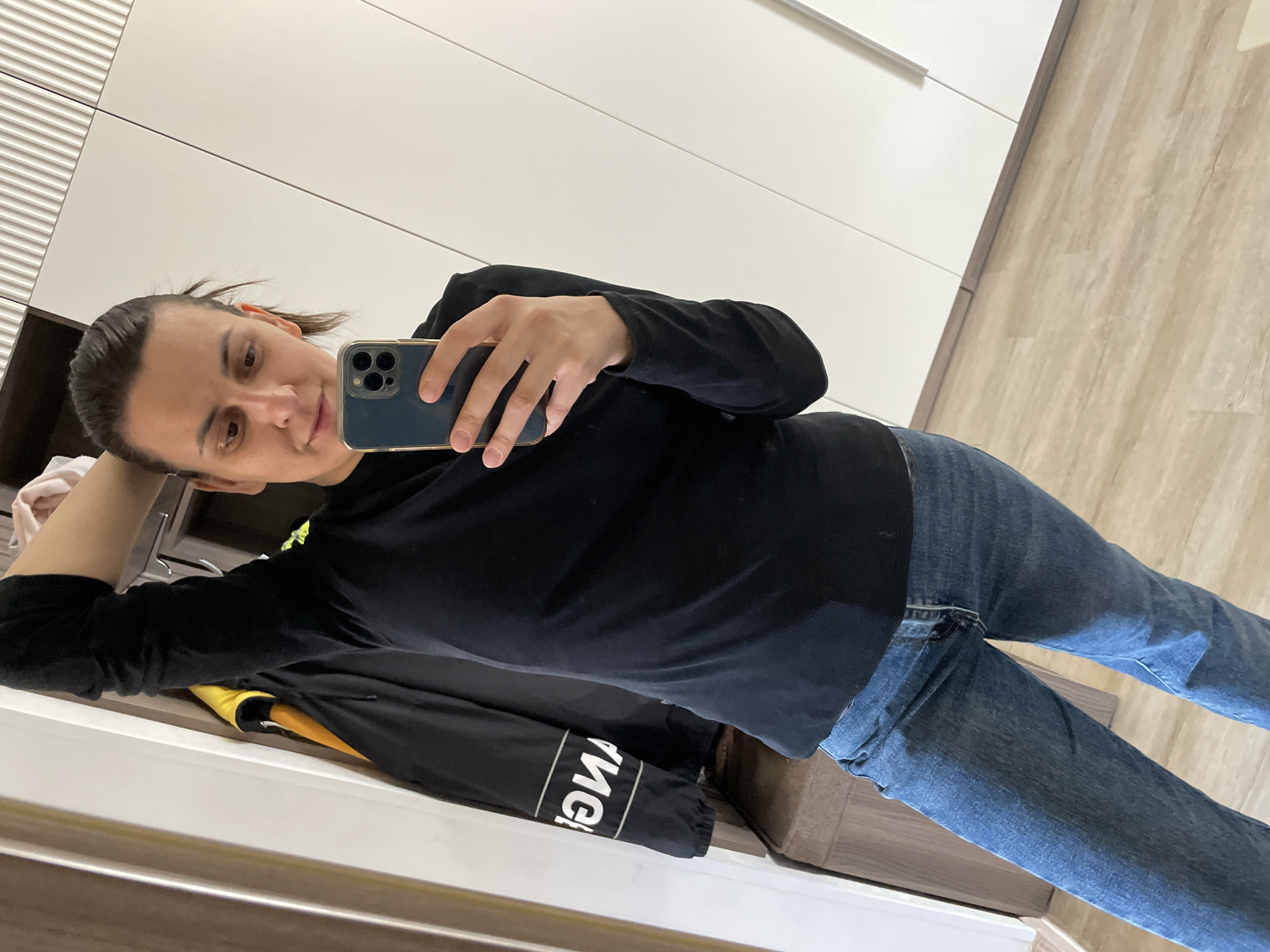 A person with shoulder-length dark hair is taking a selfie indoors. They are wearing a black long-sleeved shirt and blue jeans. The person's left hand is behind their head, and their right hand is holding a smartphone. A black jacket with white lettering is lying on a surface. The background includes white cabinets and light-colored flooring. The location is Unknown, Unknown.