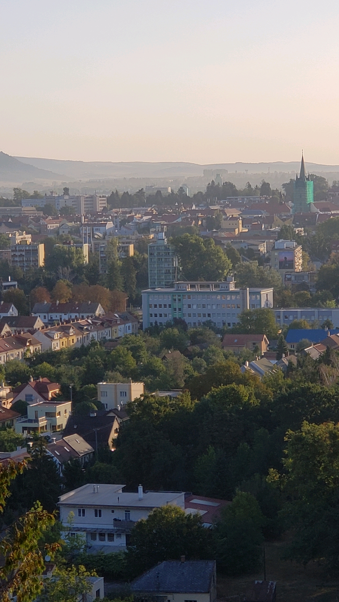 An aerial view captures a dense urban landscape during daylight, likely in the morning or late afternoon due to the soft lighting. The scene is dominated by a mix of residential buildings, featuring red-tiled roofs and varied architectural styles, interspersed with trees. In the background, a cluster of taller, modern buildings are visible, including a multi-story light-colored structure with many windows and a taller, pale blue building. To the right of the frame, a distinct green-covered church steeple rises above the rooftops. The city extends towards a hazy, distant horizon with rolling hills, suggesting a location like Prešov, Slovakia, given the architectural style and landscape. There are no people visible in the image.