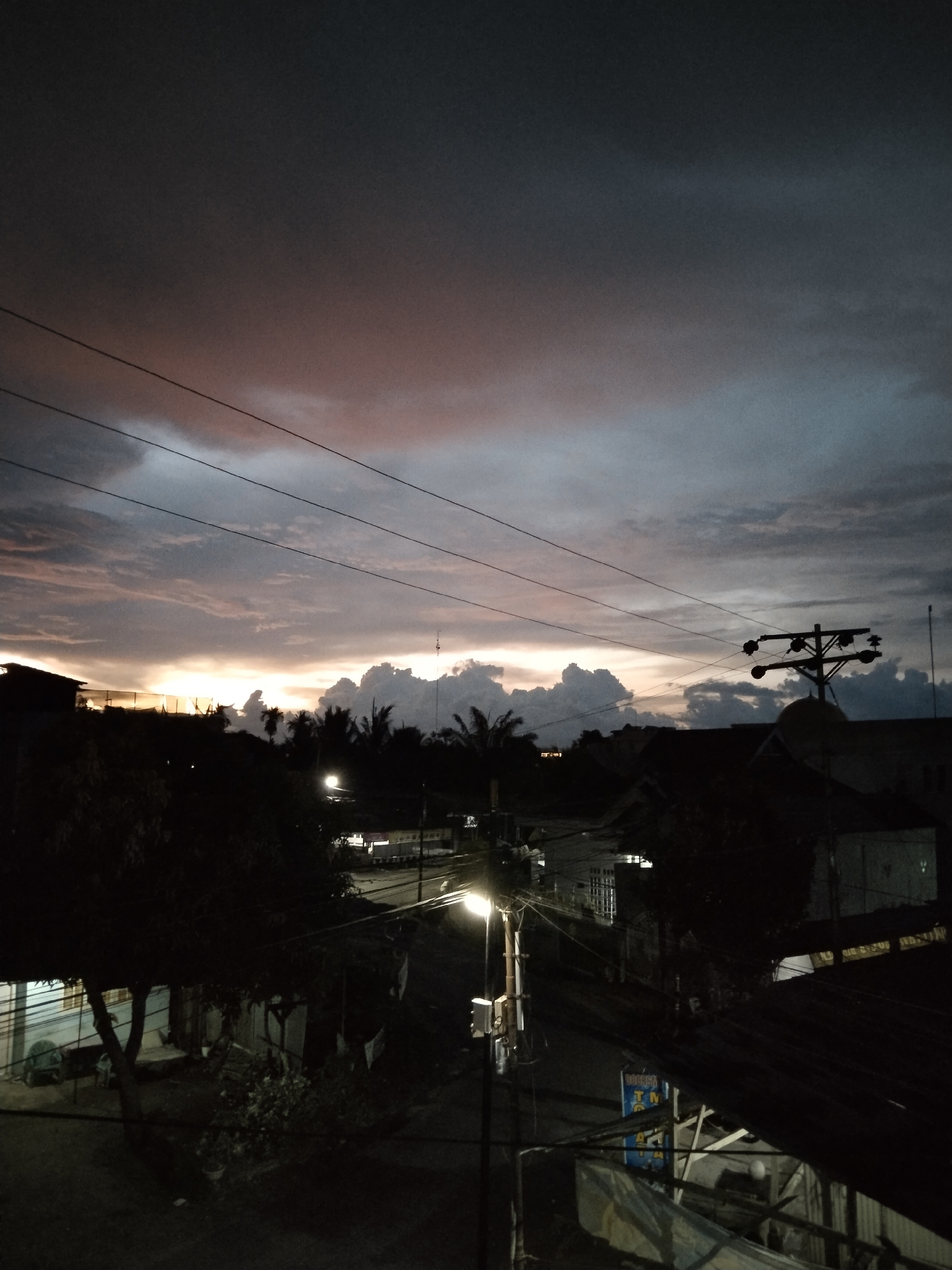 This image shows a street view in Bireuenmeunasahdayah, Indonesia, at dusk. Overhead, the sky is a mix of dark storm clouds tinged with pink and yellow light from the setting sun. In the foreground, there is a network of power lines running across the sky. Below, the street is illuminated by a single street lamp, its light casting a stark glow. The surrounding buildings are dark, with some vague details visible. Trees line the street, silhouetted against the evening sky. There are a few signs and structures visible, partially obscured by shadows and the darkness.
