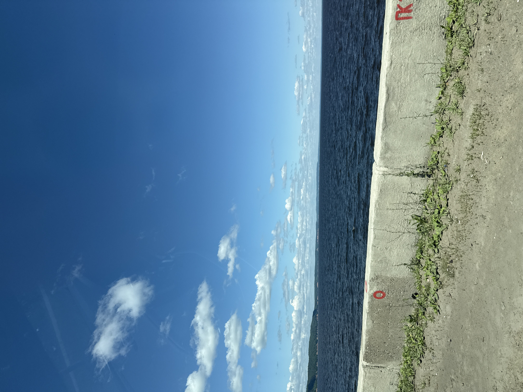 This is a medium shot taken from a slightly elevated perspective, looking out over a body of water. The image is oriented vertically. On the right side of the frame is a concrete embankment, with sparse green vegetation growing along its edge. The concrete itself has some red markings on it, including a symbol that appears to be a stylized "K" and a circular red mark. The embankment slopes down towards a dark, rippling body of water that occupies the right half of the image. The water stretches towards the horizon, where it meets a distant, hazy landmass that appears to be forested. The sky above is a vibrant, clear blue with scattered white clouds, some of which are wispy and others more substantial. The bright blue sky and the presence of clouds suggest it is daytime, likely mid-morning or afternoon, with fair weather. The overall impression is one of a serene, natural landscape with some human infrastructure. No people are visible in the image.